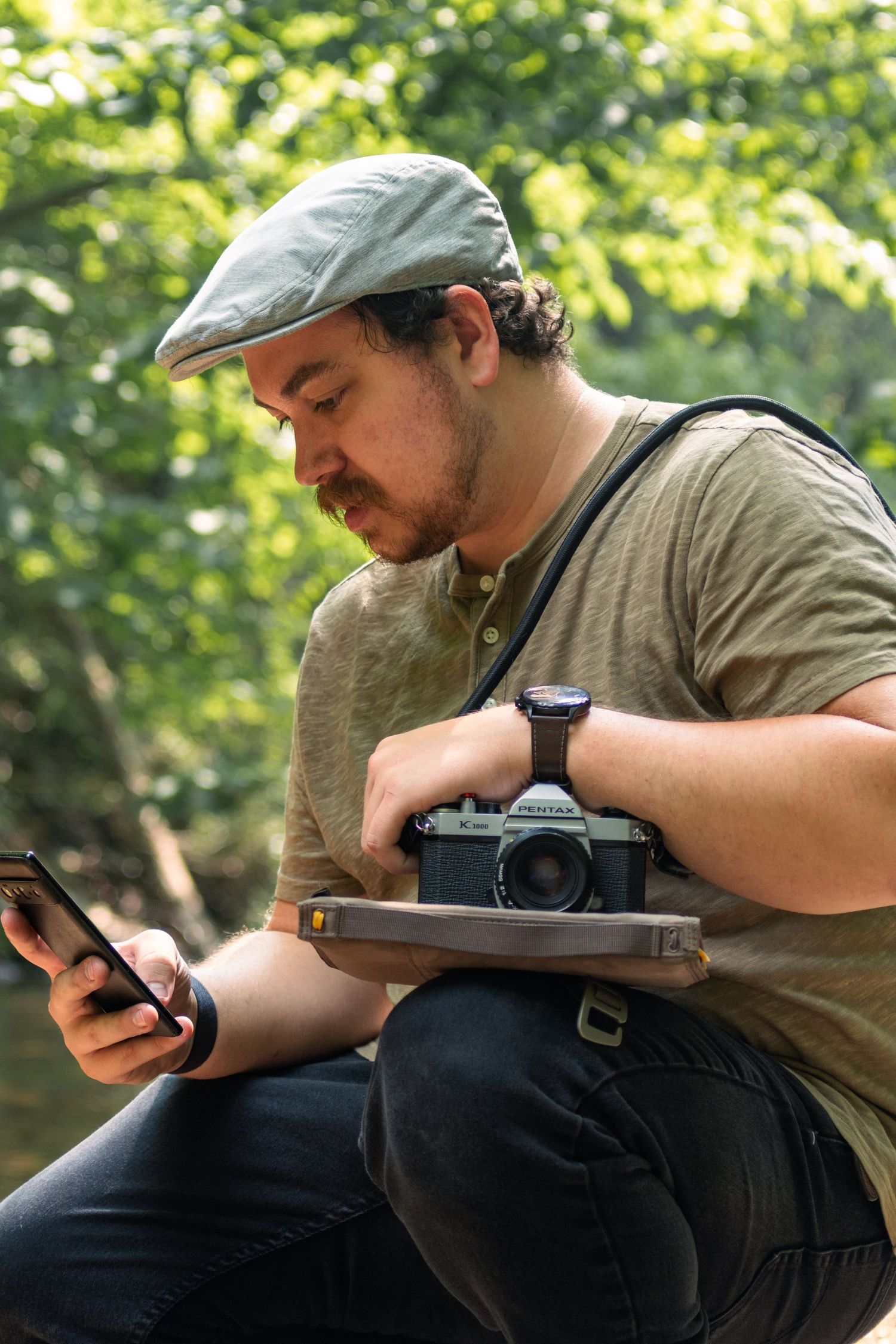 Photographer in newsboy cap checks phone while holding vintage camera outdoors.