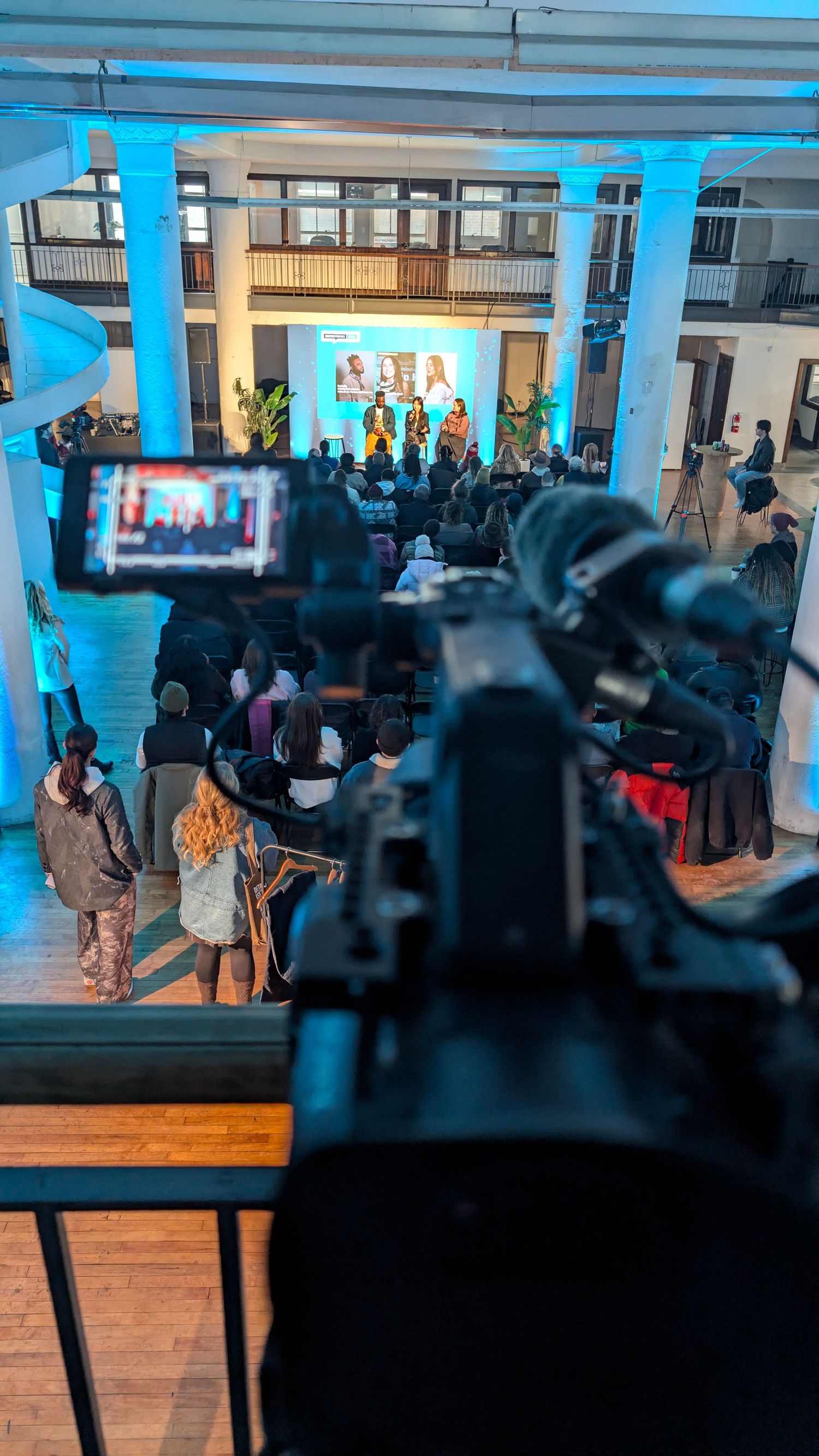 Professional video camera records a live performance with blue stage lighting and audience in an indoor venue.
