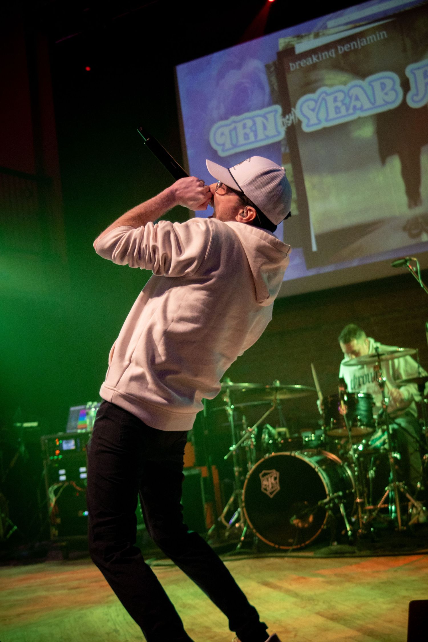 A performer in a pink hoodie dances energetically on stage with green lighting and drum equipment visible in the background.