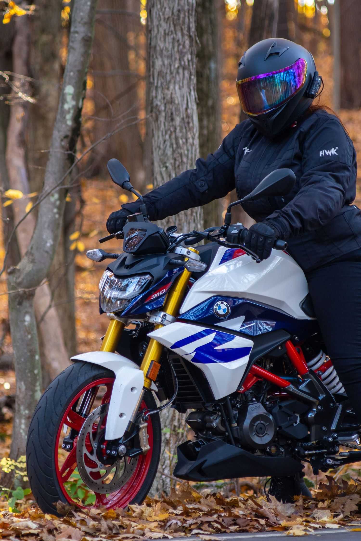 BMW G310R motorcycle rider in black gear and helmet poses in autumn forest with fallen leaves at sunset.