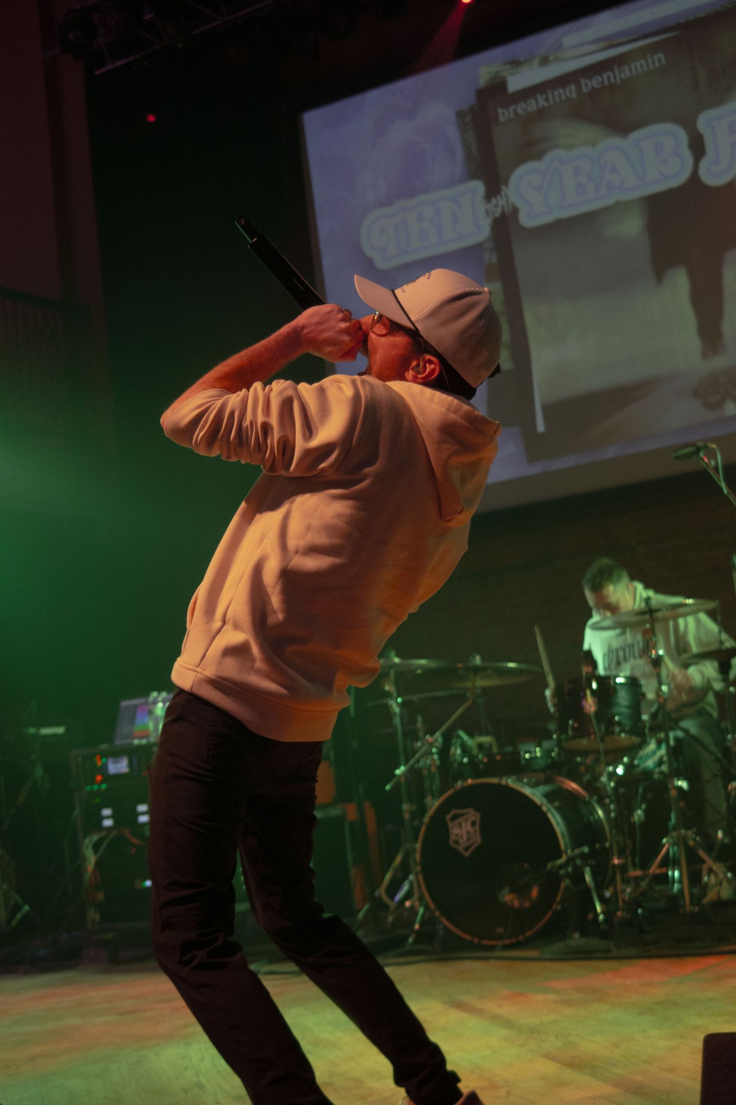 Singer performing on stage with microphone, wearing white hoodie and cap, with drummer and projection screen visible behind.