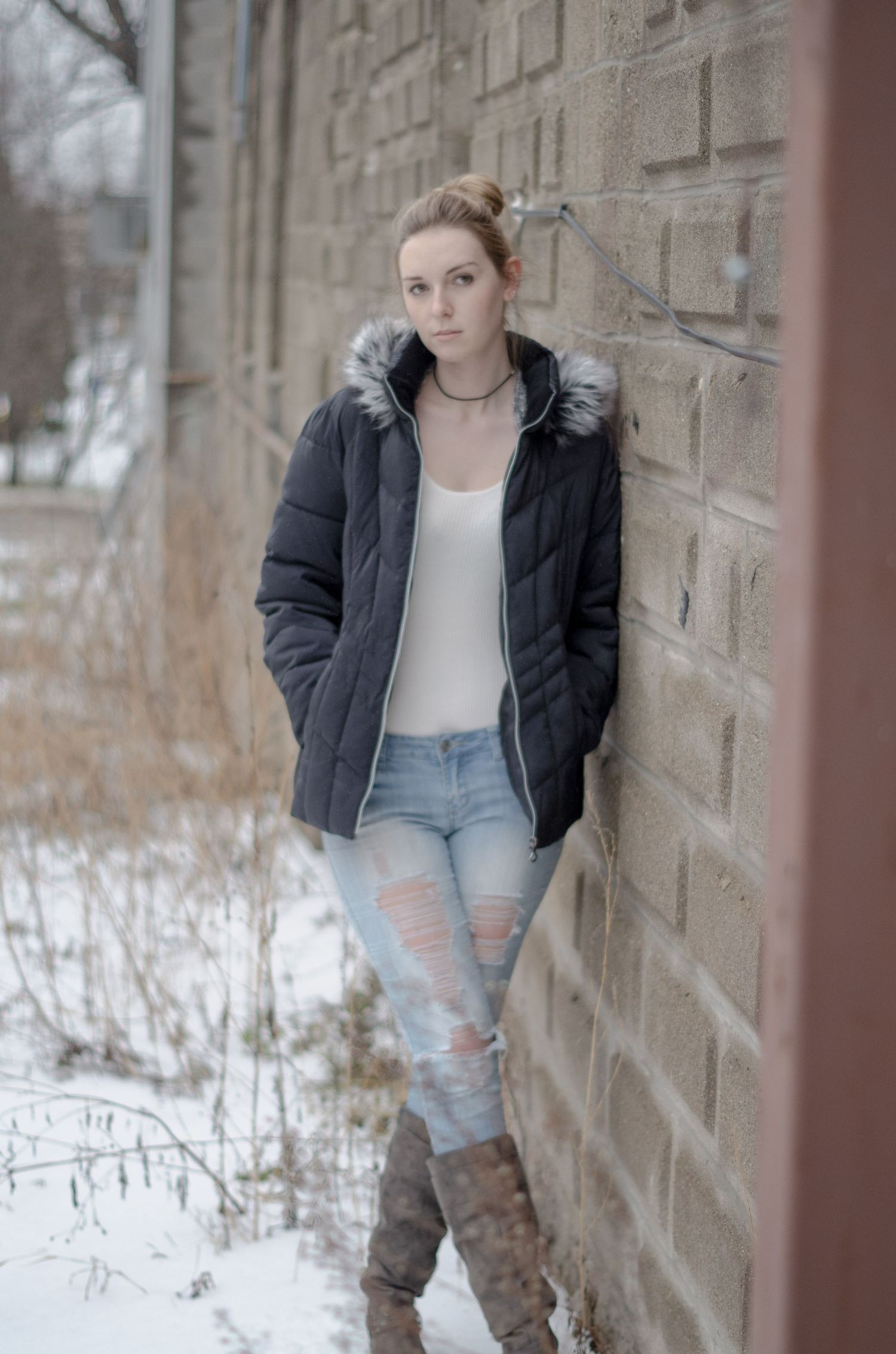 Winter fashion portrait against brick wall showing winter jacket, distressed jeans and boots in snowy setting.