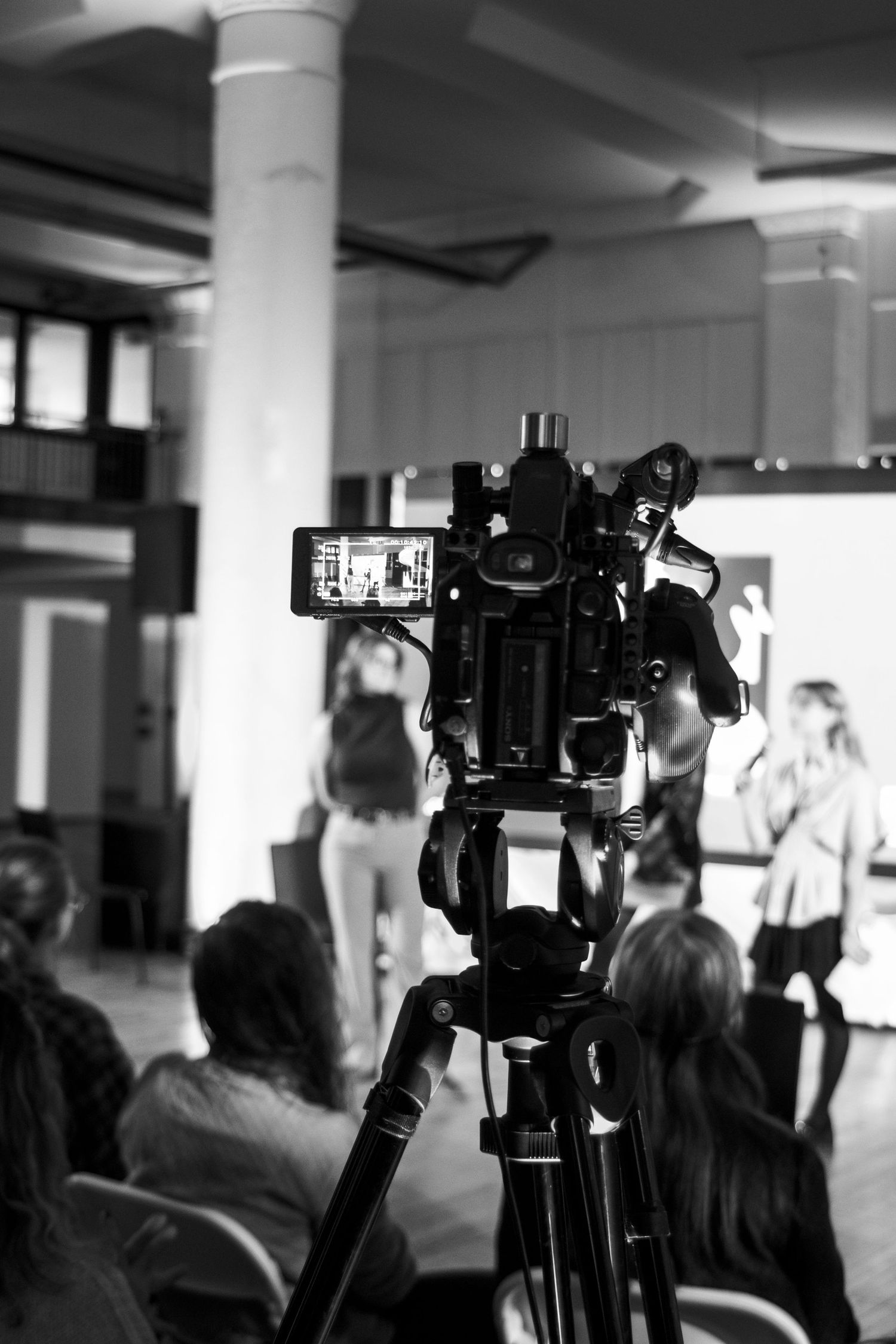 Professional video camera on tripod set up at indoor event with people visible in background in black and white photography.