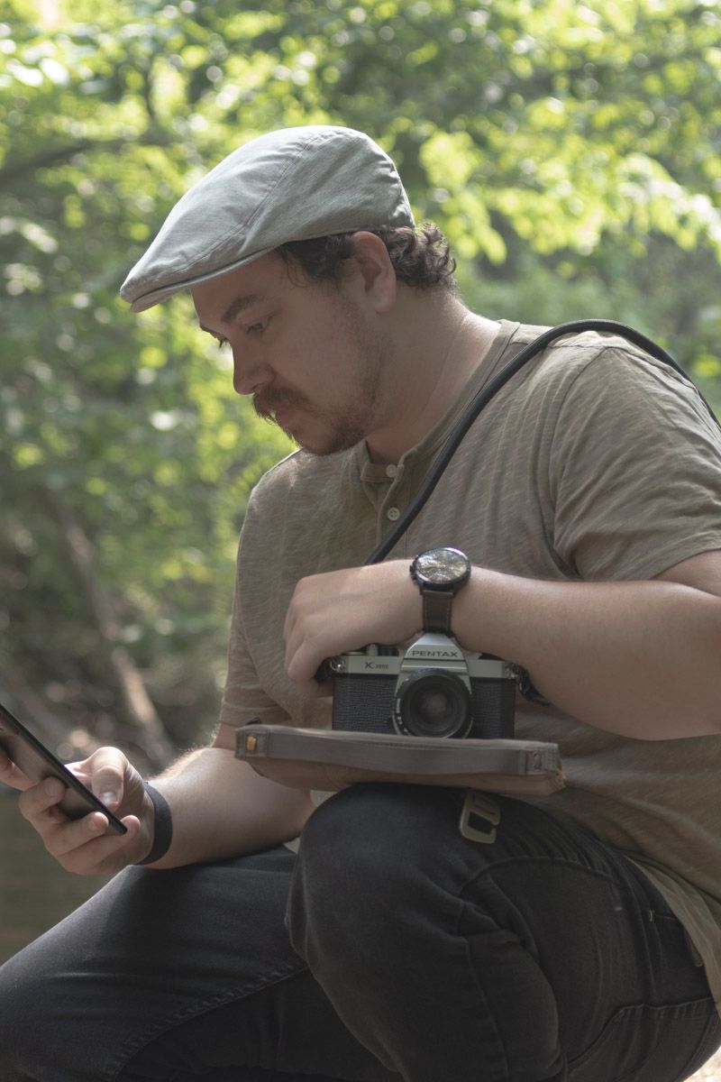 Photographer in flat cap checking phone while holding vintage camera.