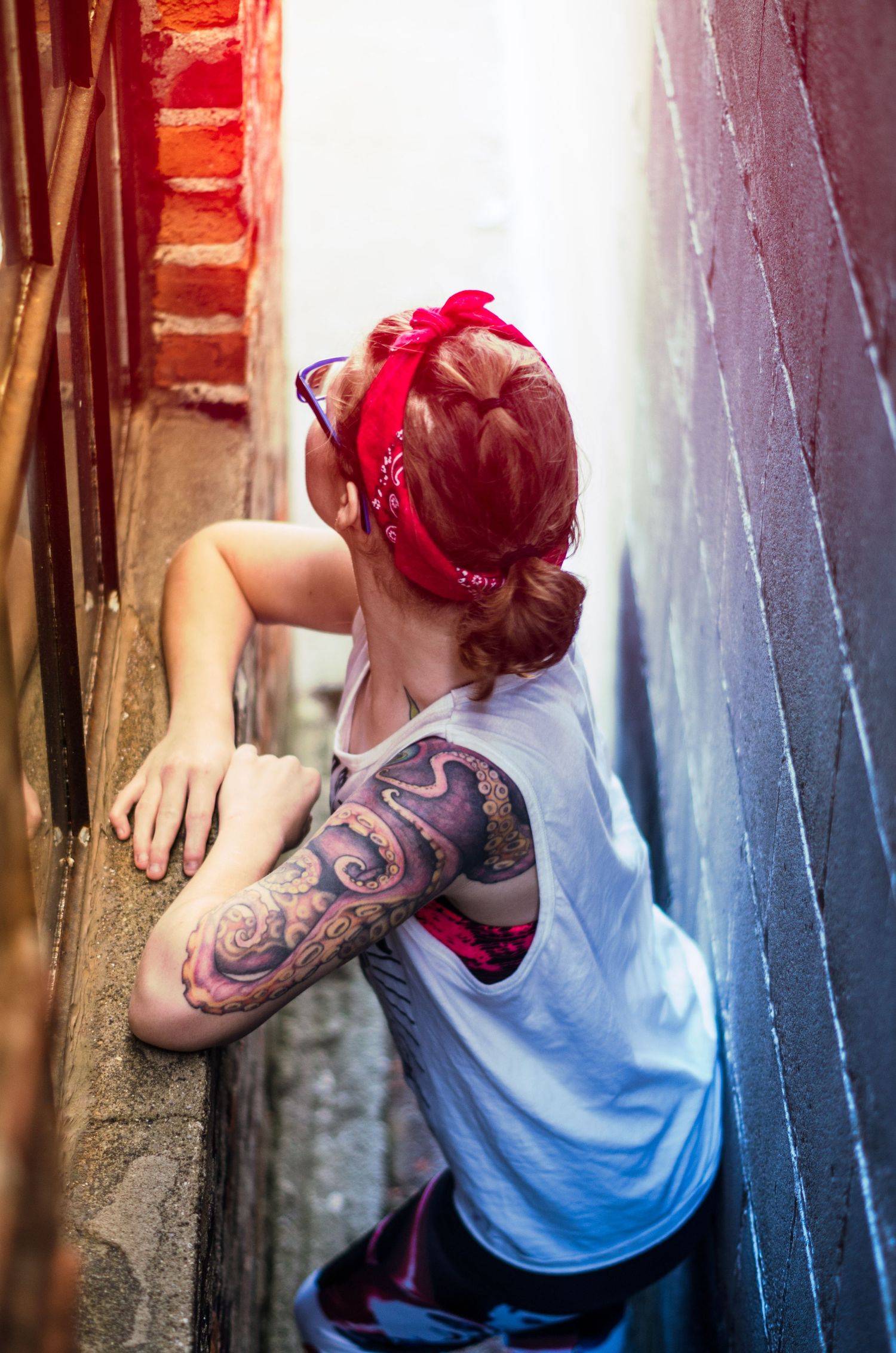 Person with red bandana and arm tattoo leaning against brick wall in artistic pose.