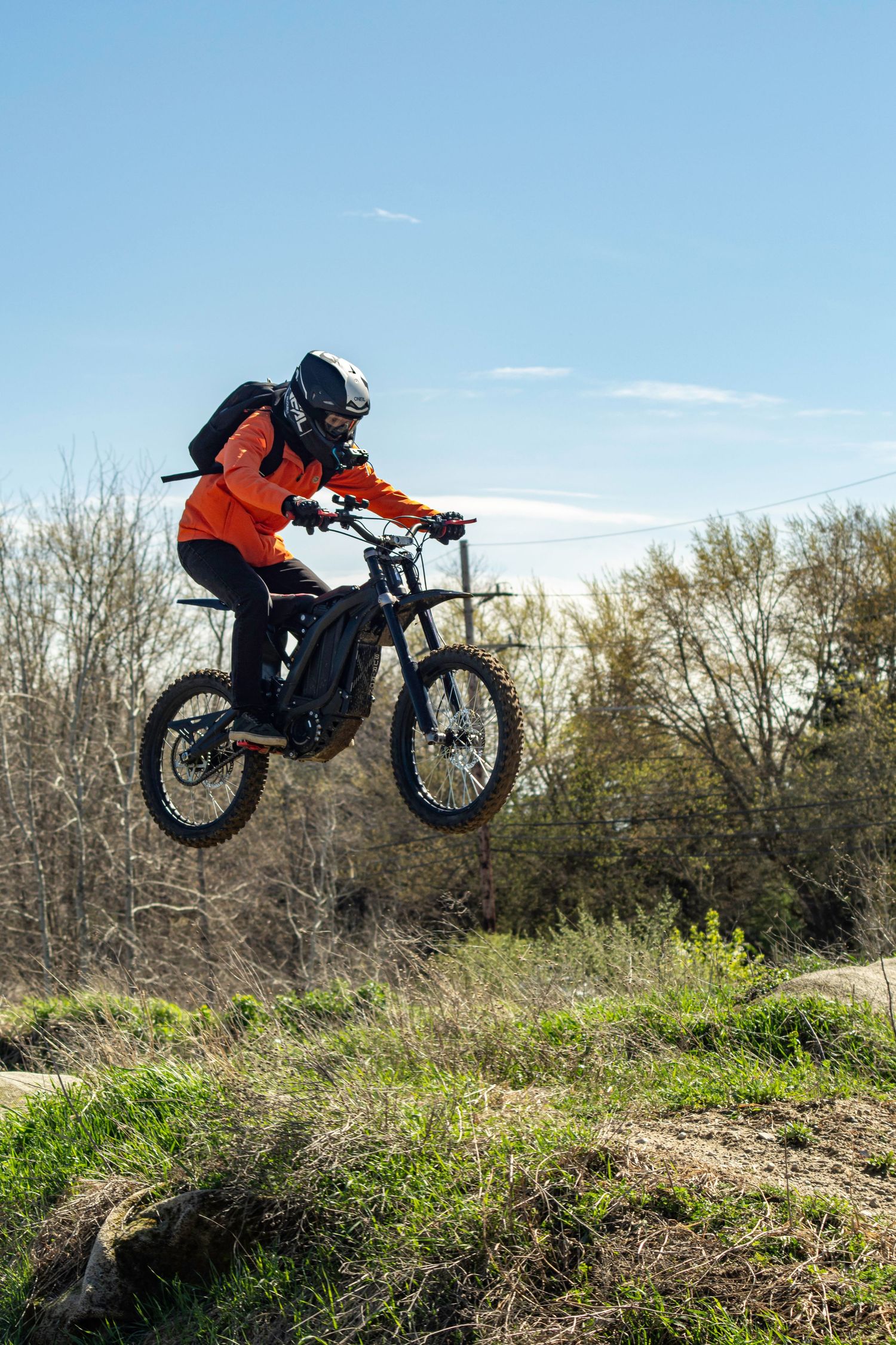 Mountain biker in orange jacket performing aerial jump off dirt mound against blue sky.