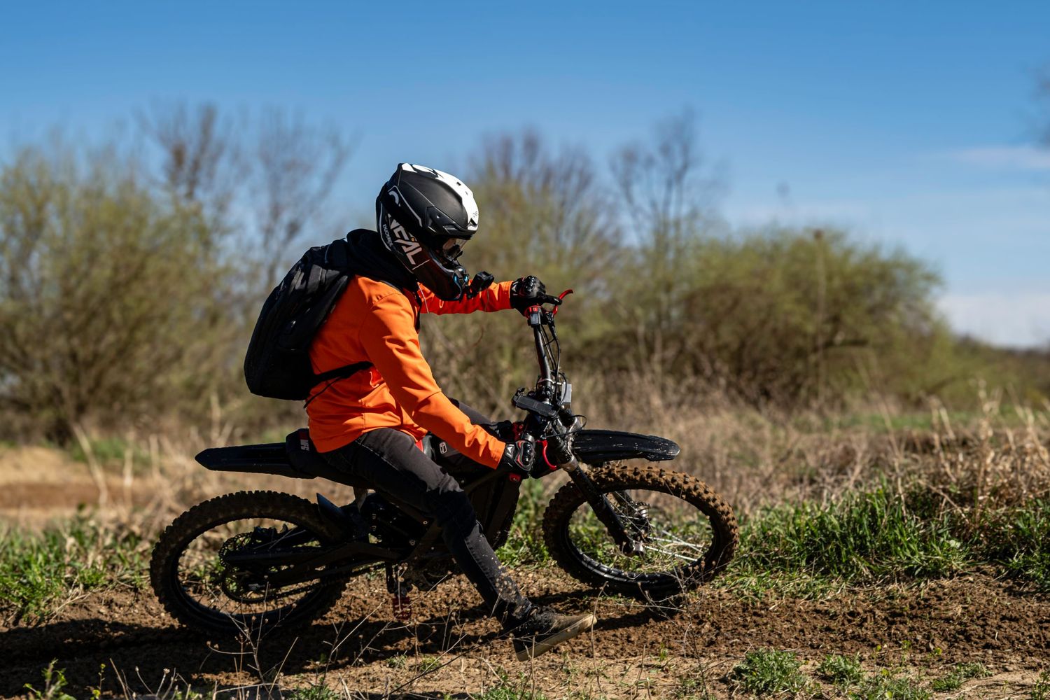Dirt bike rider in orange jacket and helmet riding through a rural dirt trail.