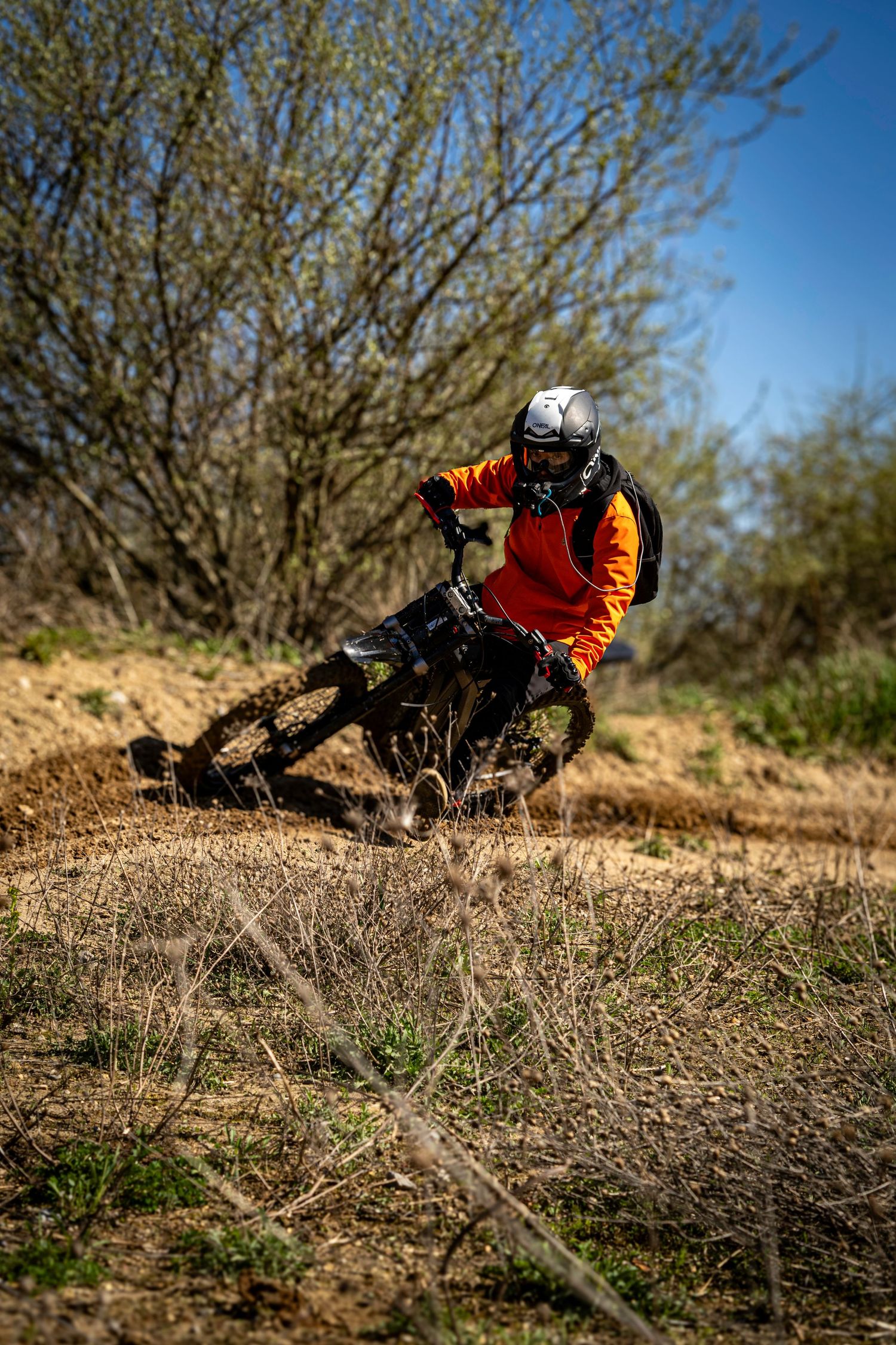 Dirt bike rider in orange jersey navigating a dusty turn on an off-road trail during a sunny afternoon.