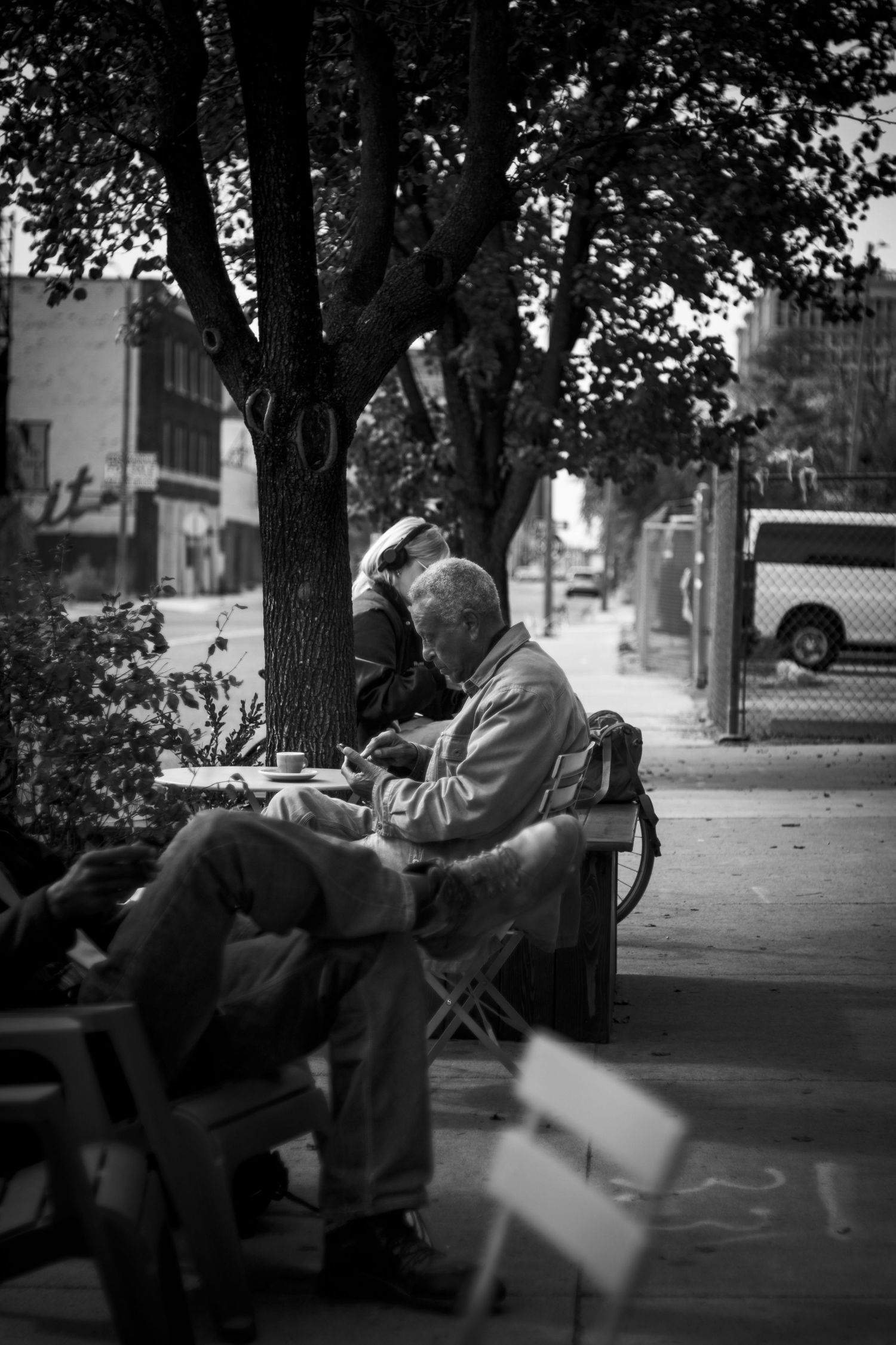 Black and white photo of an elderly man reading on a bench beside a tree on a city sidewalk.