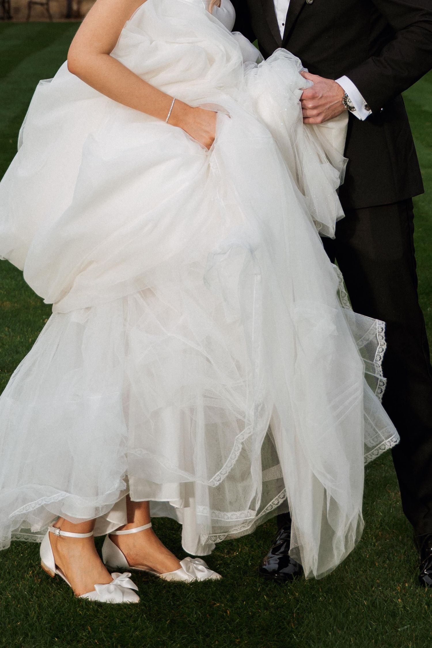Close-up shots of a bride's feet in white heels peeking out from under layers of tulle wedding dress on grass.