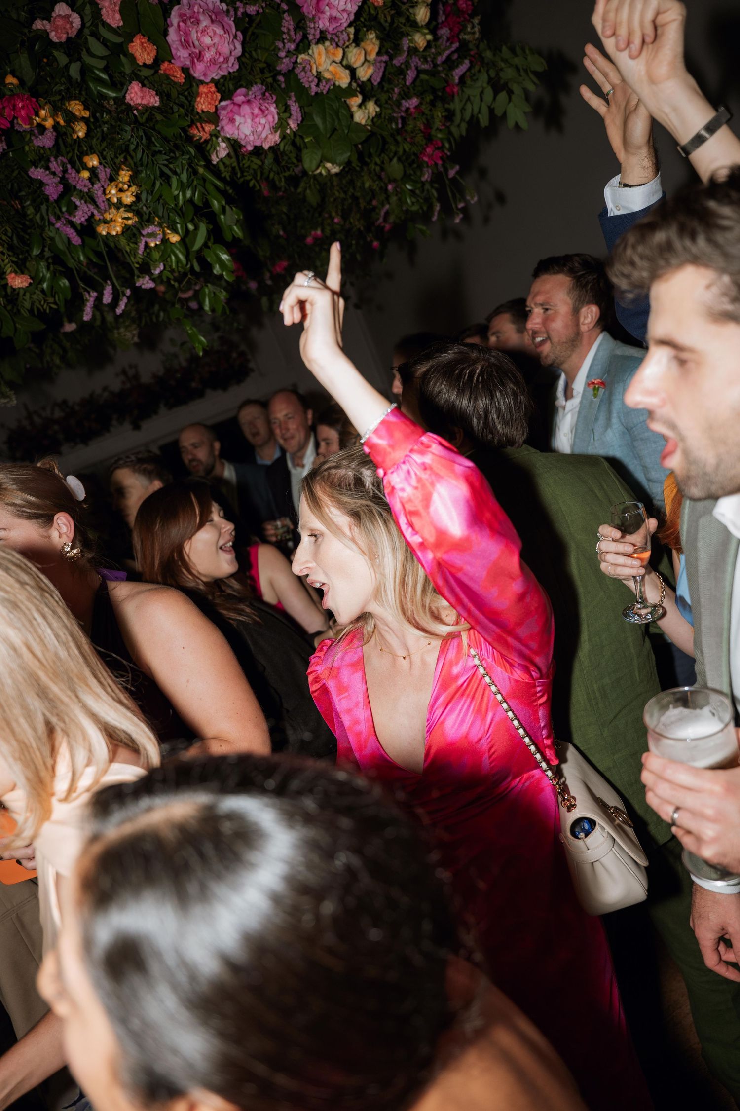 A crowd of people dancing and celebrating at a nighttime party with pink lighting and floral decorations.