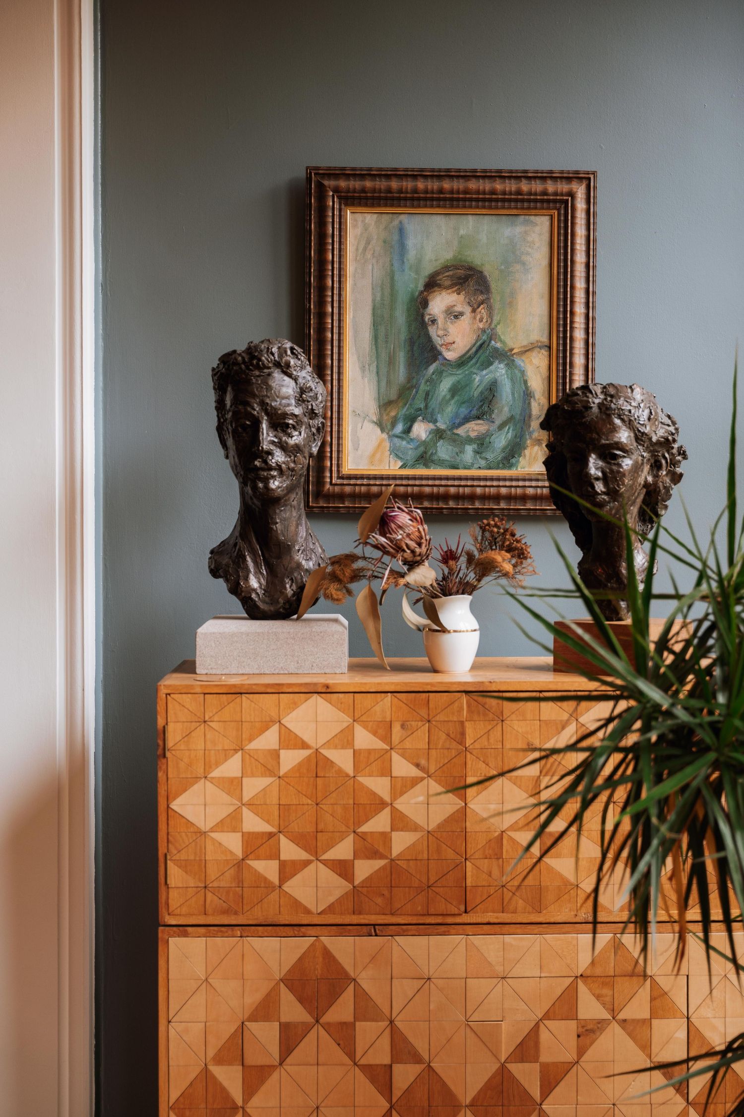 Bronze busts displayed on wooden geometric cabinet with framed artwork and plants in an interior setting.