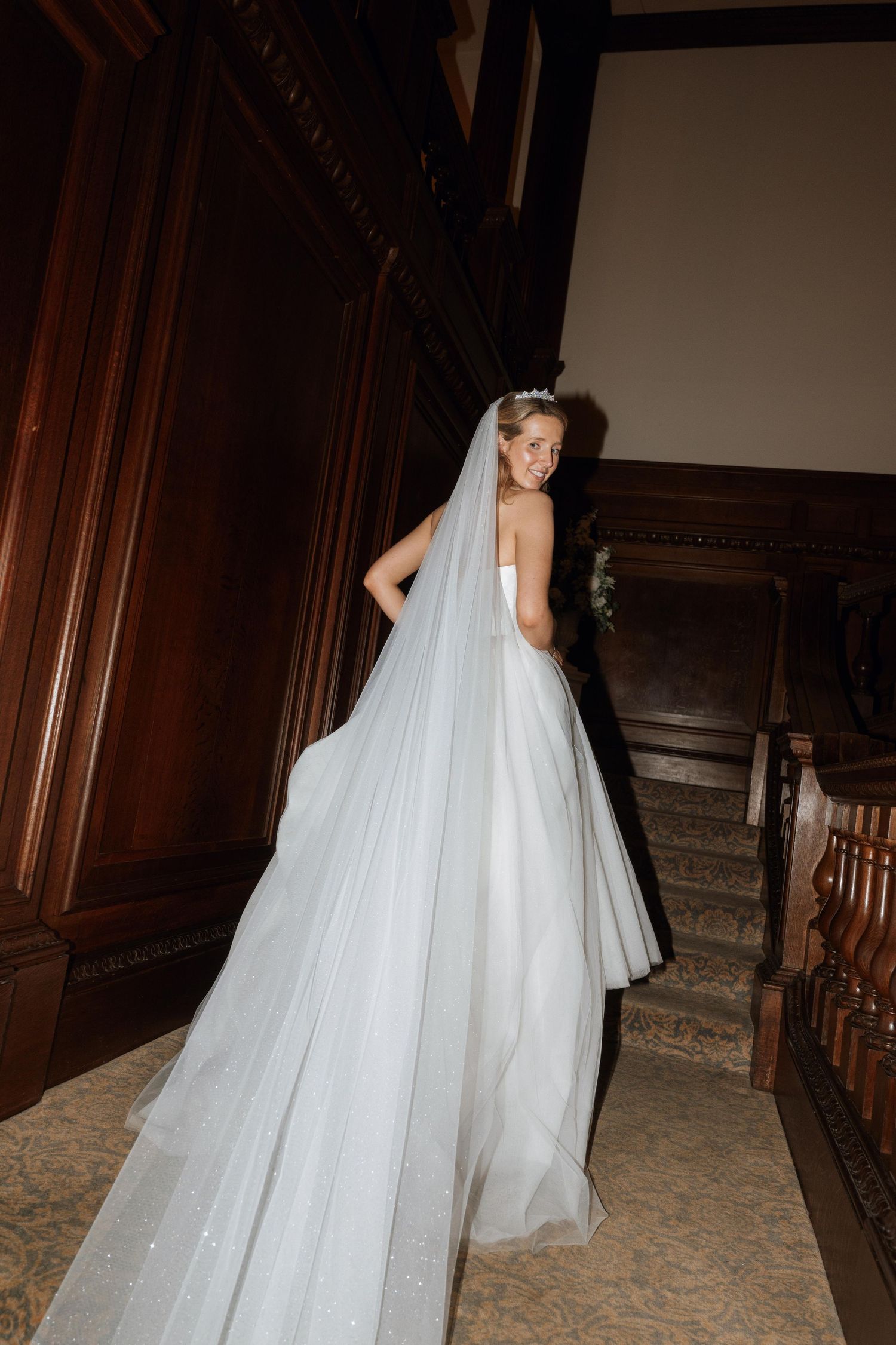 A bride in a flowing white wedding gown poses gracefully on a staircase in an elegant indoor setting.