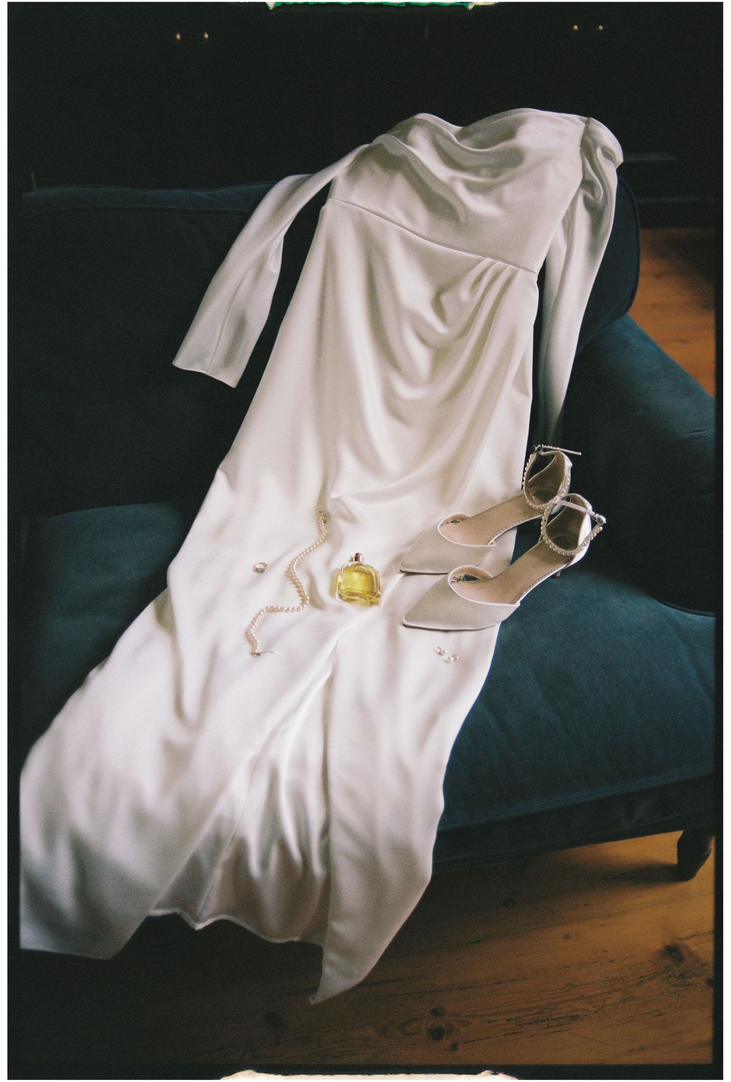 A white elegant dress laid out on a dark couch with beige heels and a small perfume bottle.