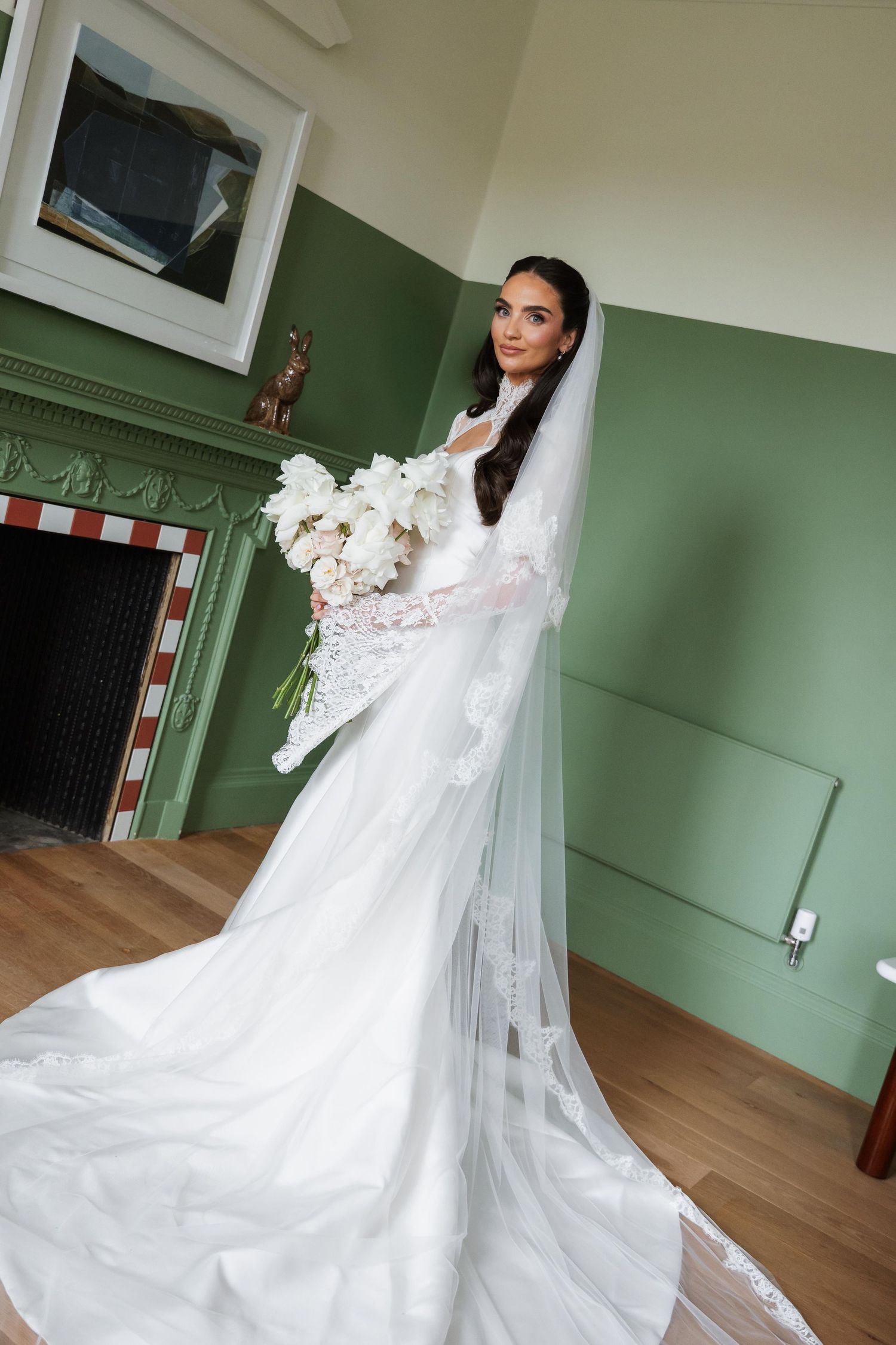 Bride in a white lace wedding gown with long veil holding a white bouquet, standing in a green room by a fireplace.