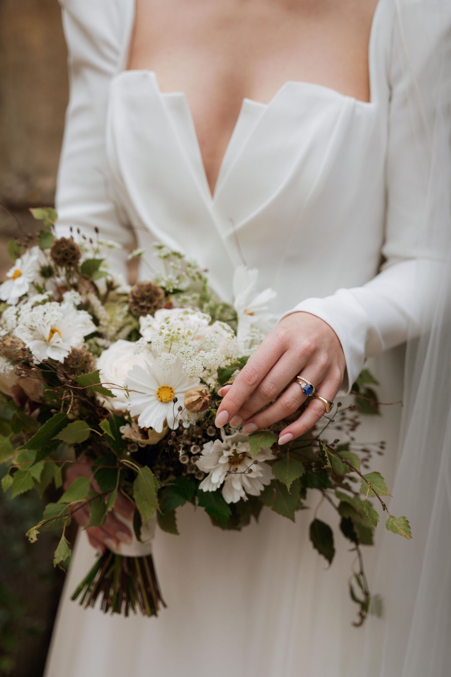 Close-up of a bridal bouquet with white flowers and greenery held by hands wearing an engagement ring and white dress.