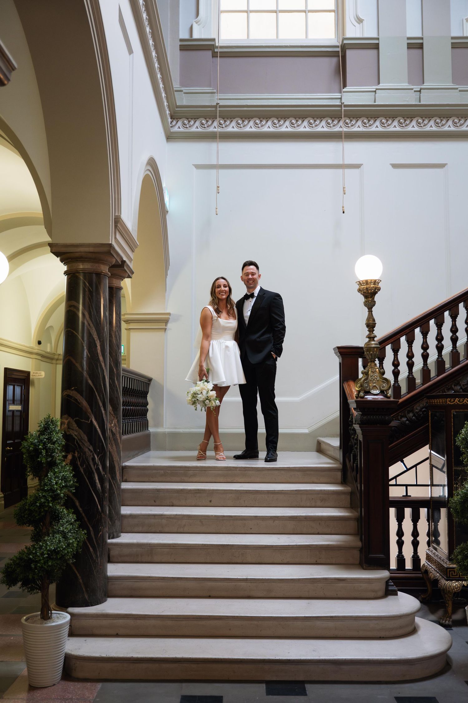 A couple stands elegantly on a grand staircase with white walls and wooden railings in a classic interior setting.