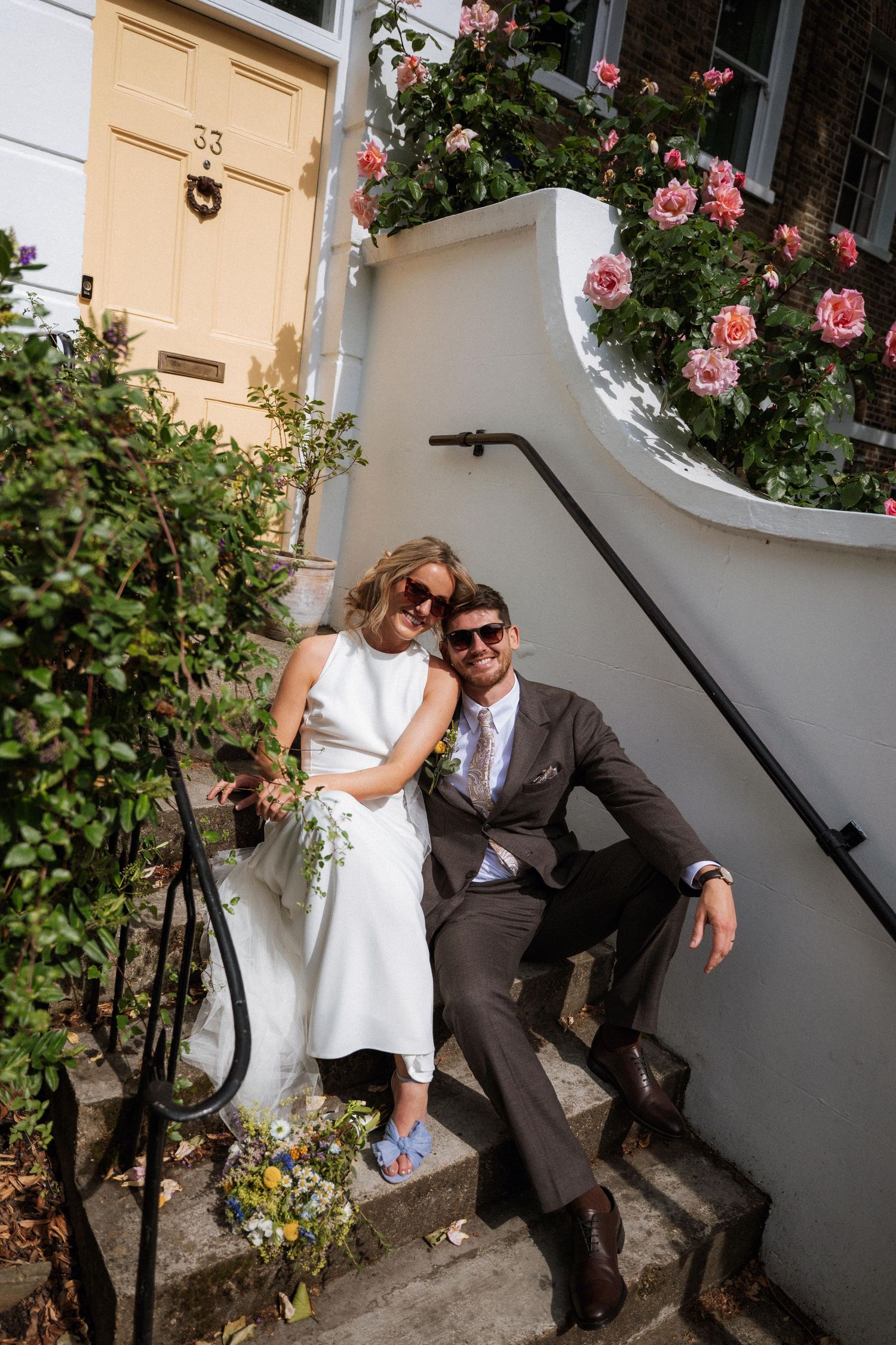 A couple shares an intimate moment on outdoor steps surrounded by pink roses and greenery.