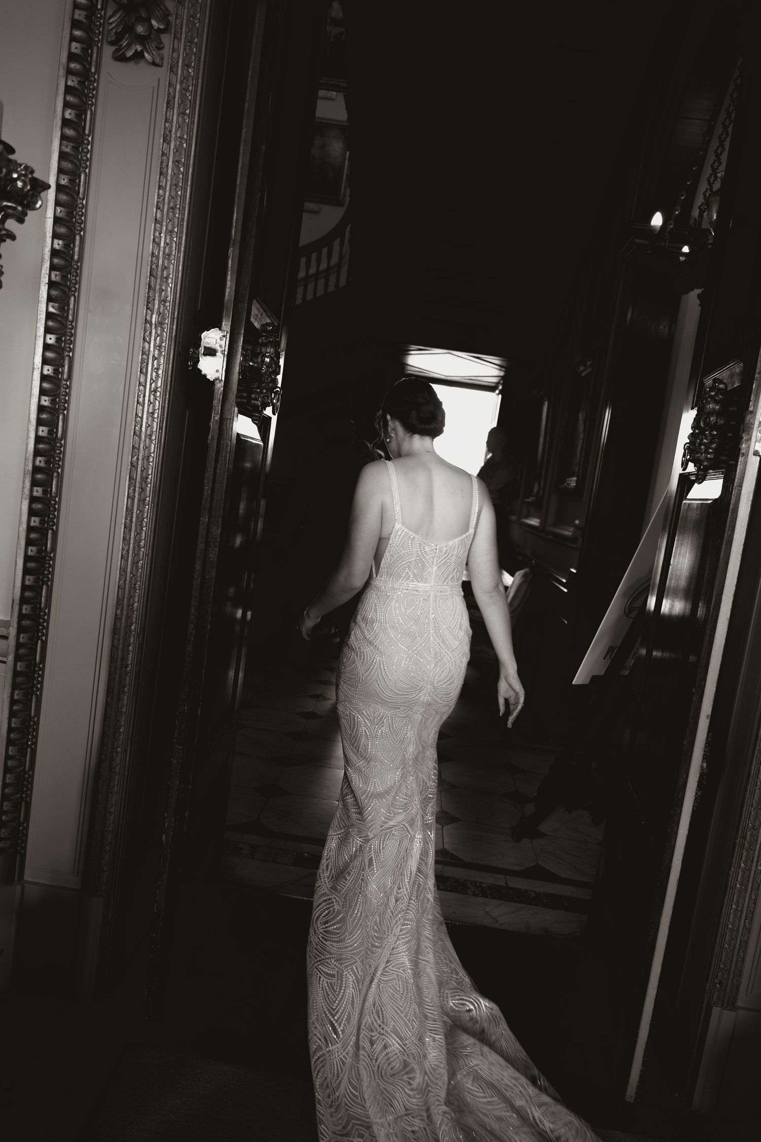 Black and white photo of a bride in a fitted lace wedding gown walking down a dark hallway.
