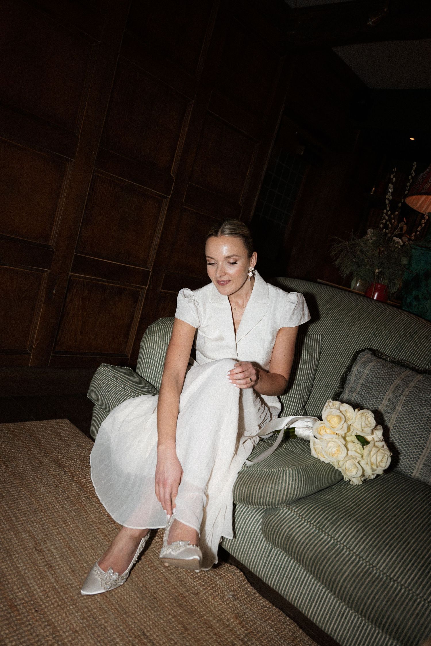 A person in an elegant white dress sits on a green sofa next to yellow roses in a dimly lit room.