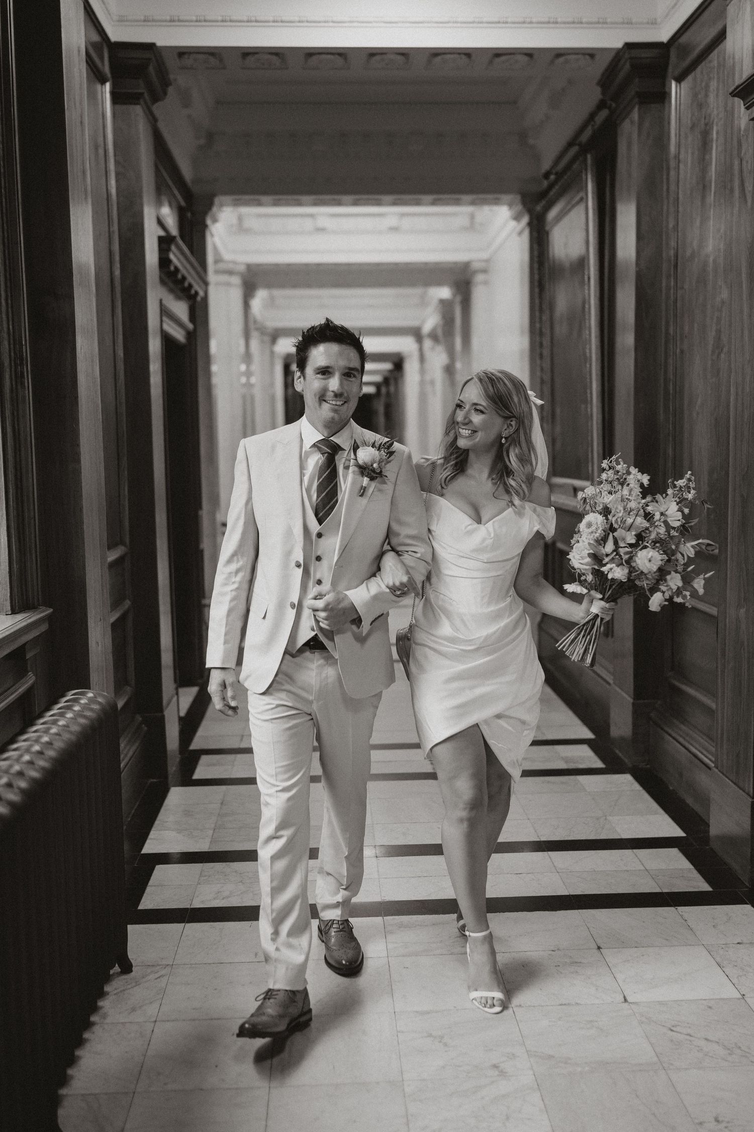 A couple in wedding attire walks playfully down an elegant hallway in a series of black and white photos.