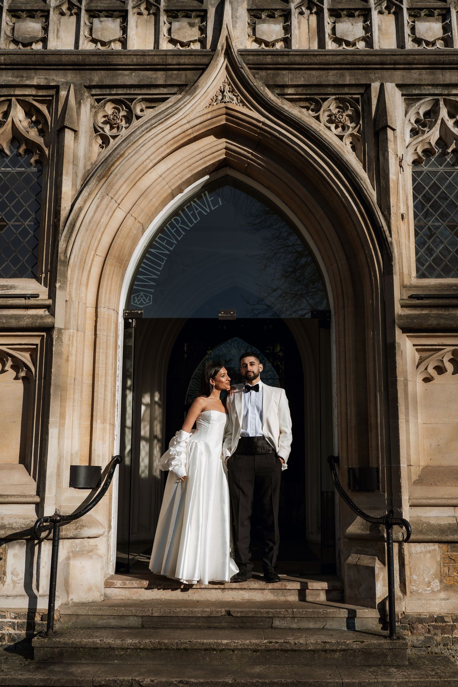 A couple in wedding attire stands in a grand Gothic church doorway with ornate stone carvings and architectural details.