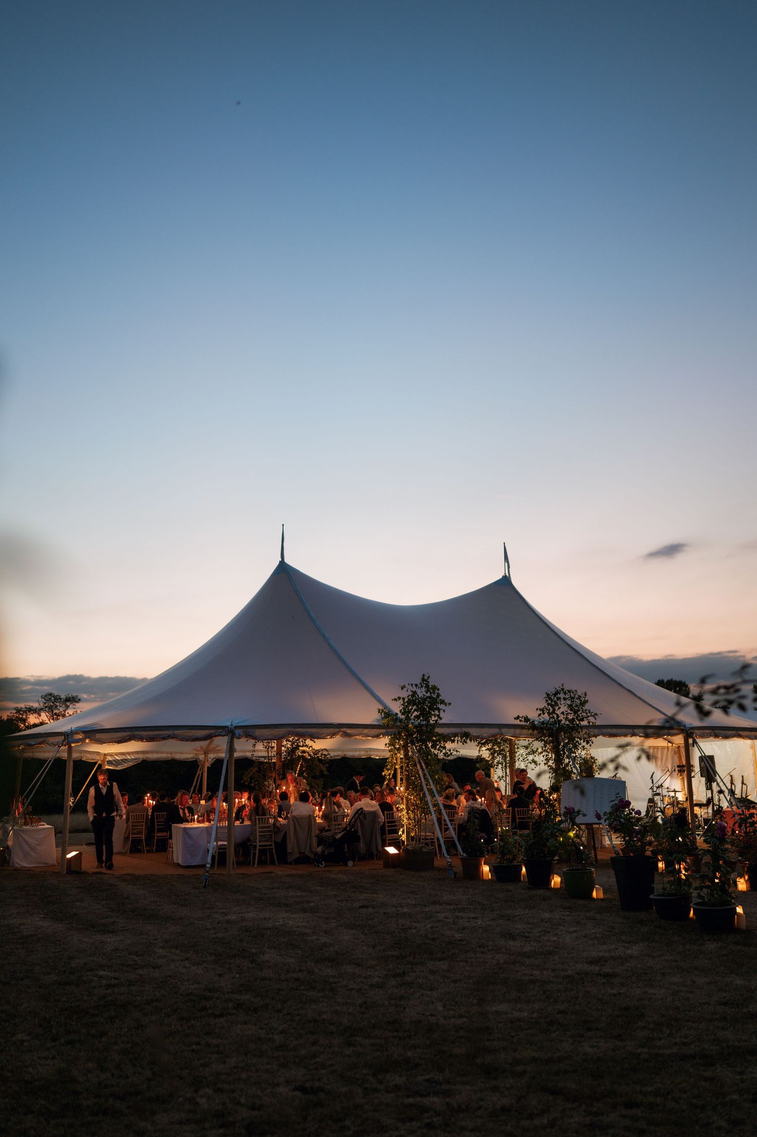 Wedding reception tent illuminated at dusk with warm lights and silhouetted guests gathered for an outdoor celebration.