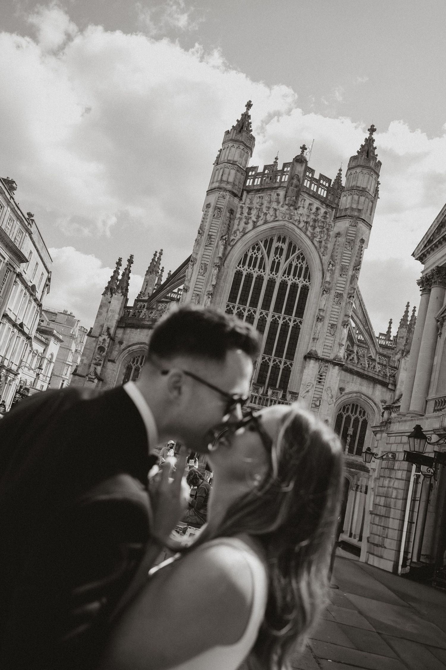 A romantic couple shares a kiss in front of the historic Gothic architecture of Bath Abbey in black and white.
