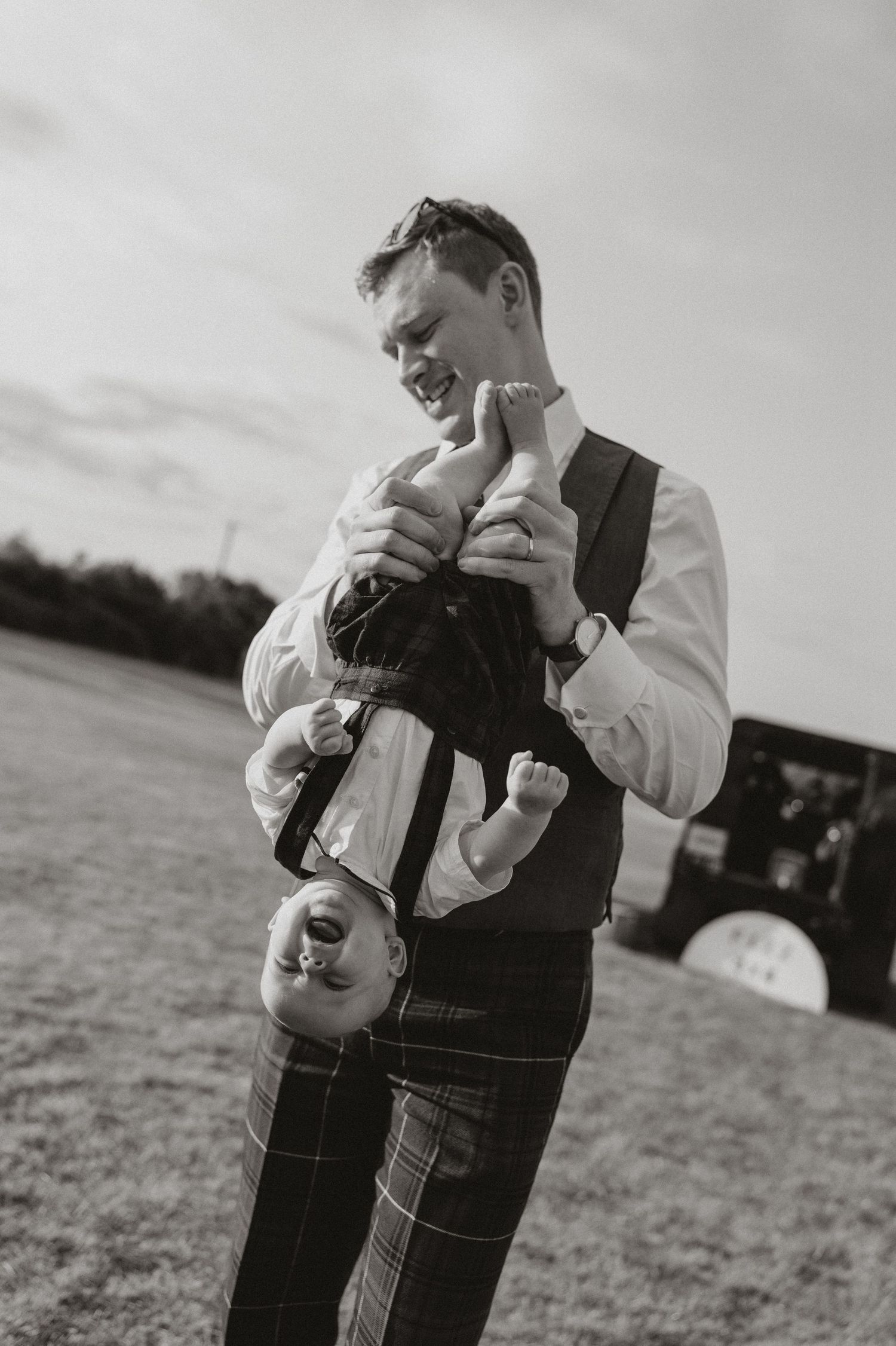 Black and white portrait of a parent in a vest holding a baby upside down playfully in an outdoor field setting.