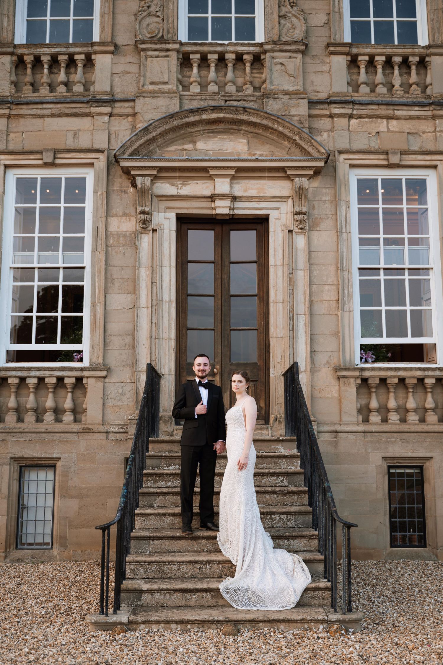 Elegant couple stands on grand stone steps of historic mansion with ornate architectural details and large windows.