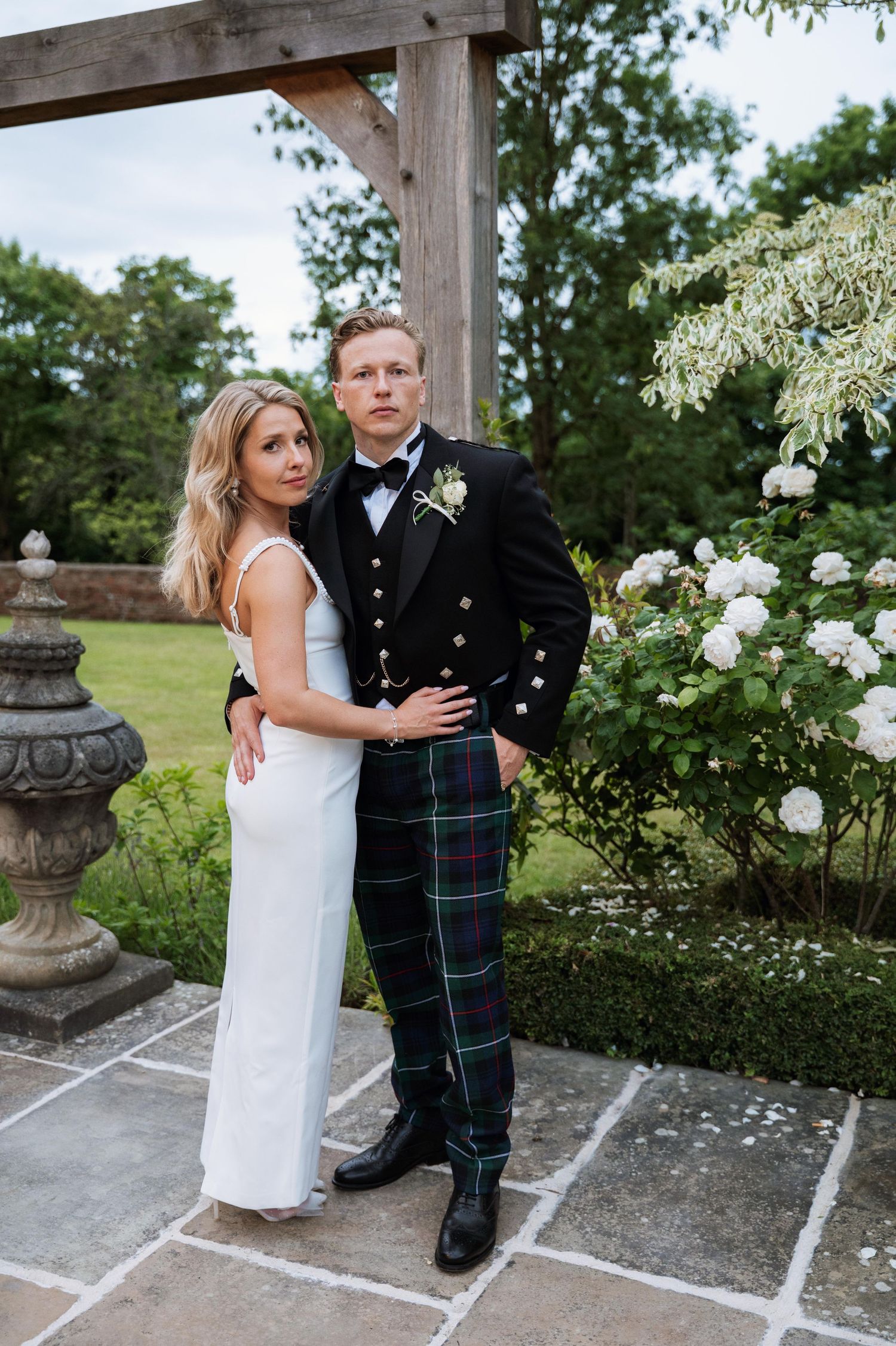 A couple in wedding attire embraces on a stone patio surrounded by lush greenery and white flowers.