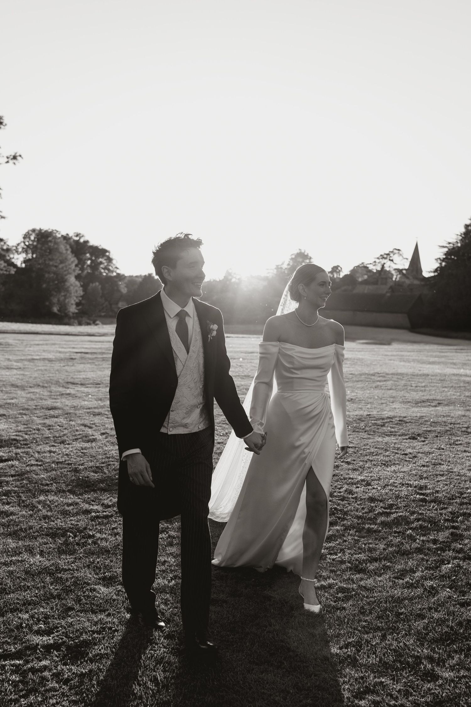 A bride and groom walk hand in hand across a sunlit lawn during their black and white wedding photoshoot.