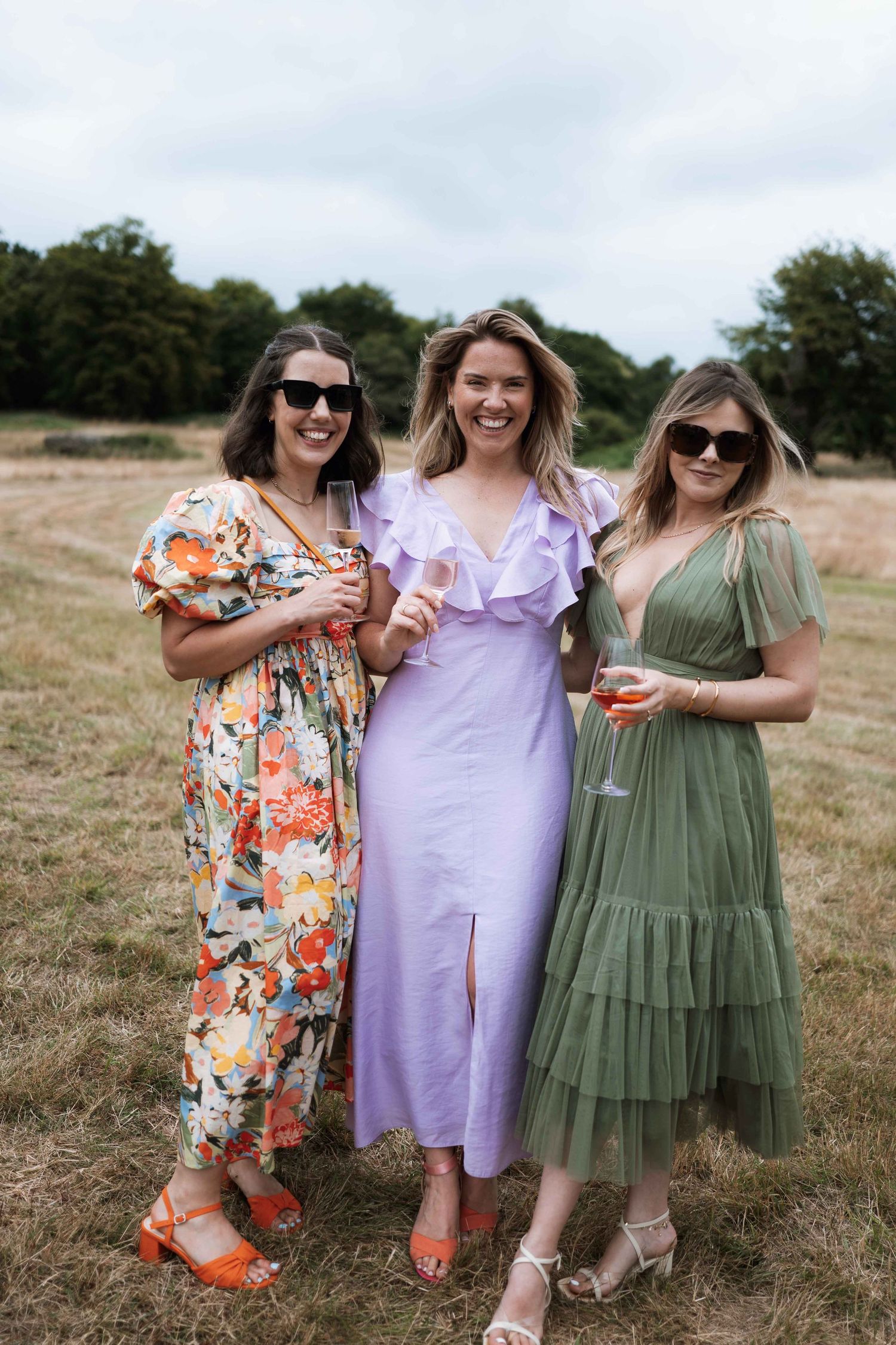 Three friends in colorful summer dresses pose together in a grassy field while smiling and enjoying each other's company.