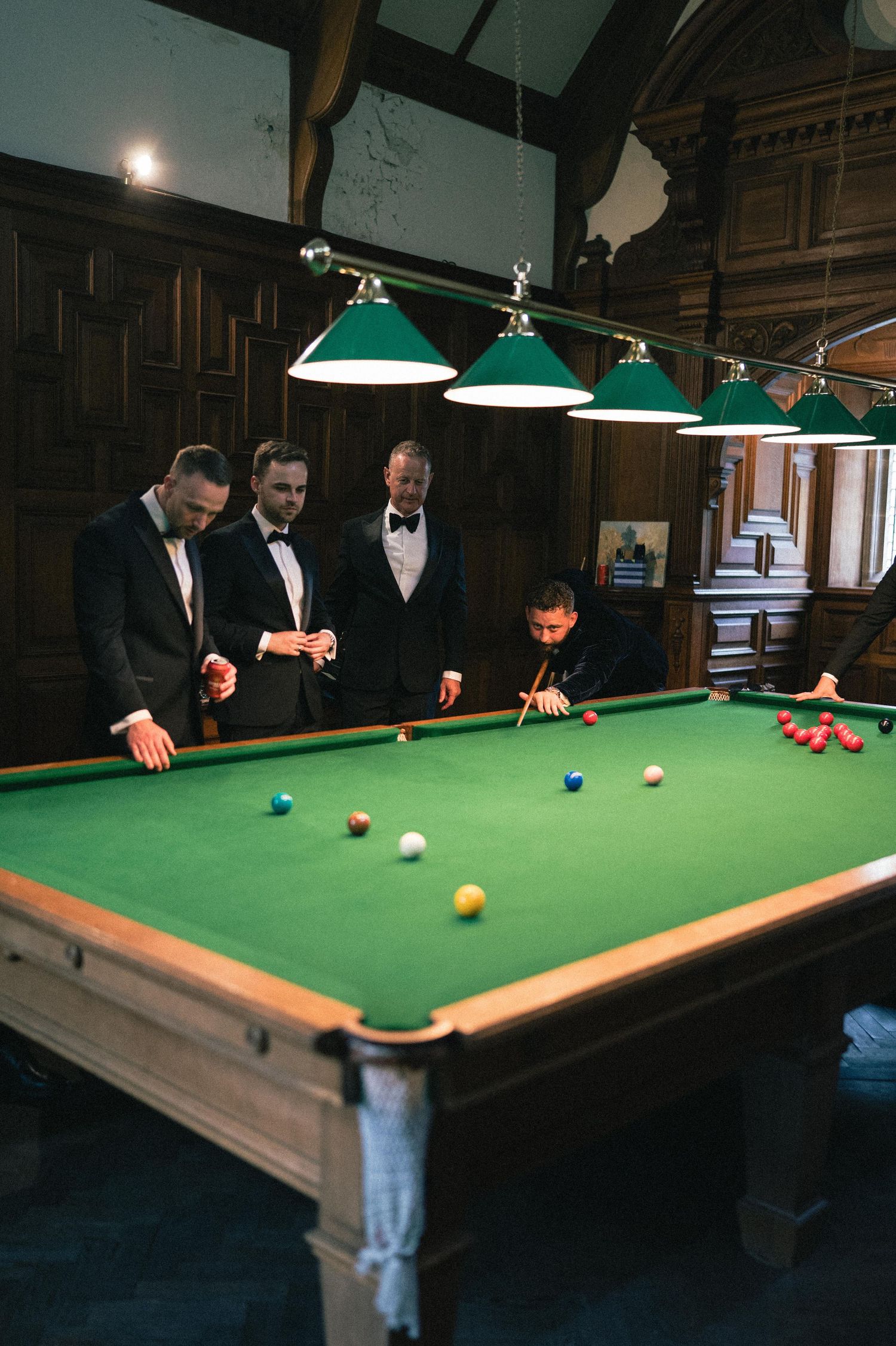 Players gather around a pool table in a dimly lit billiards room with green felt and hanging pendant lights.