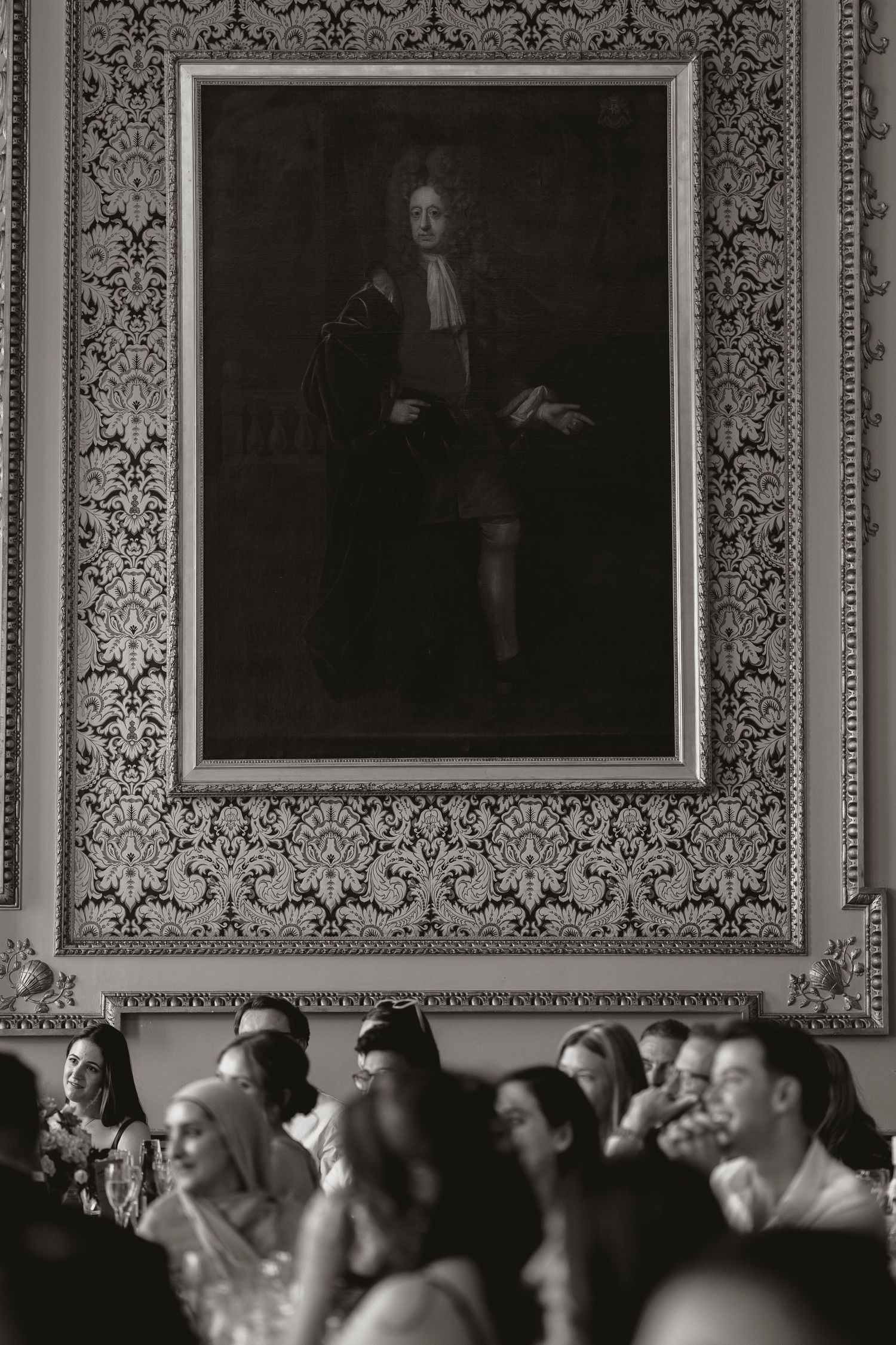 A crowd of people seated in a formal room with ornate wallpaper and framed portraits hanging on the wall.