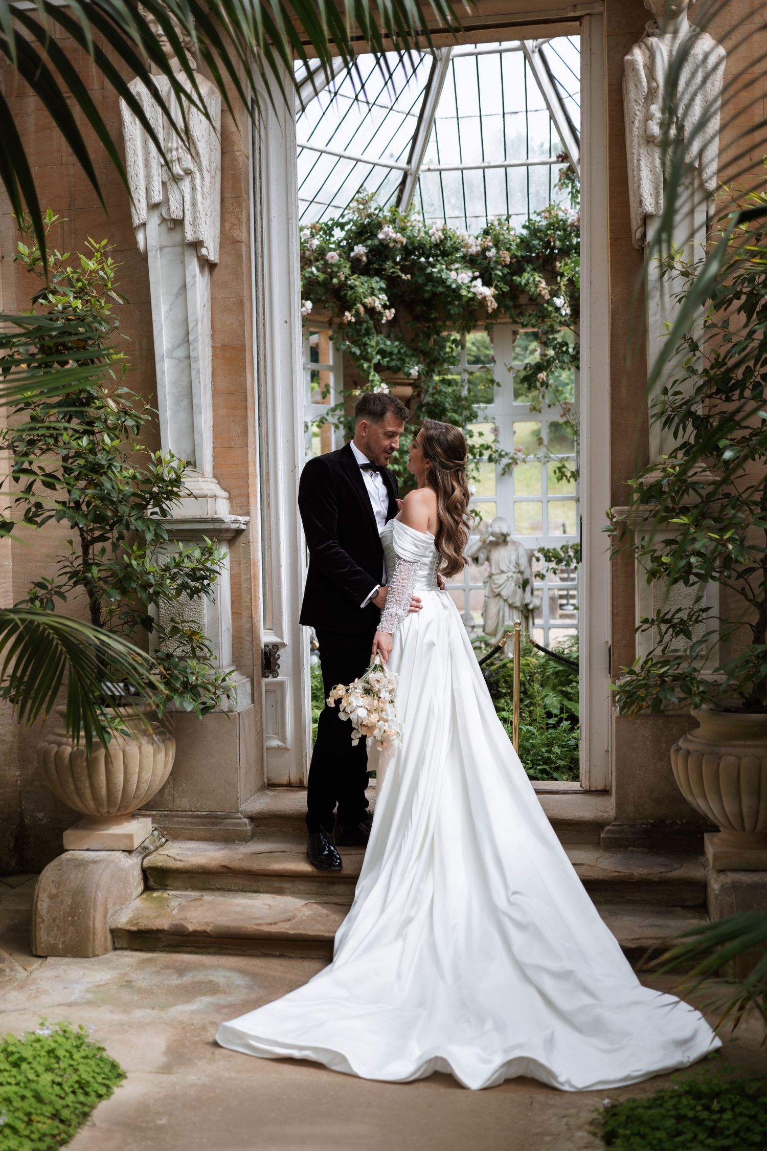 A romantic wedding photo in a greenhouse conservatory with lush greenery and white floral decor framing the elegant couple.