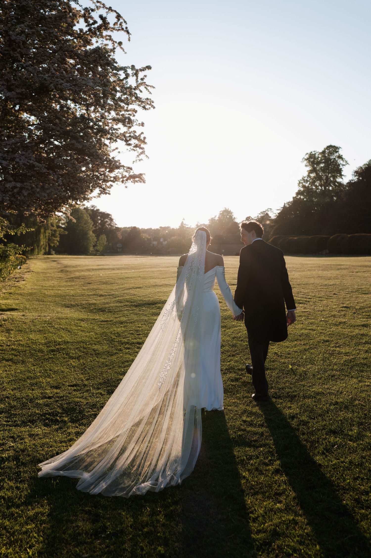 A couple in formal wedding attire walking across a sunlit grassy field with a long flowing veil trailing behind them.