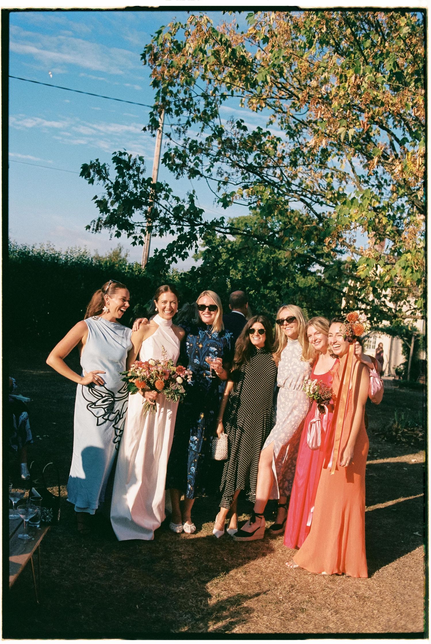 A group of friends in pastel and coral dresses gather outdoors under autumn trees during a social gathering.