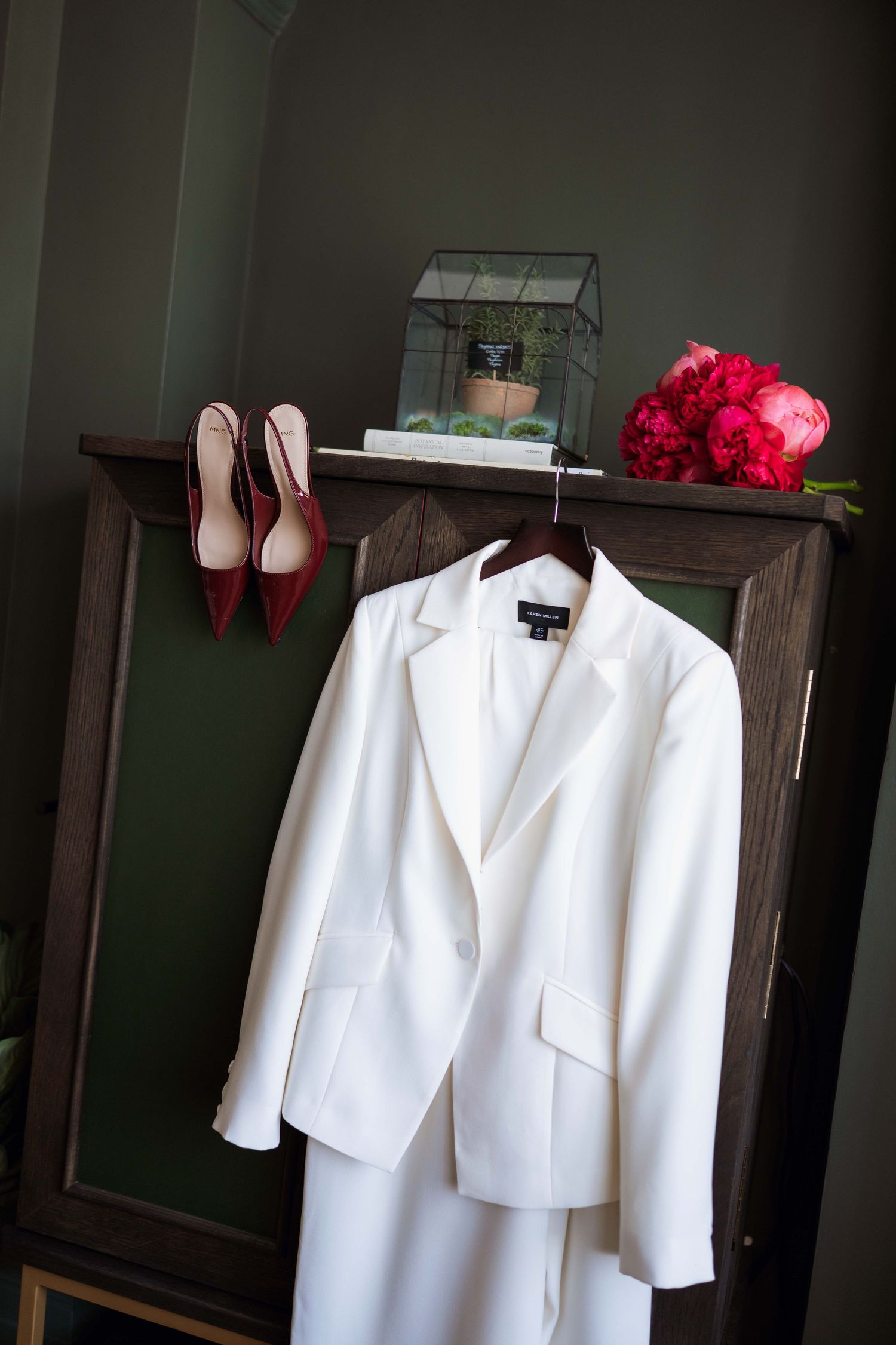 A white wedding suit jacket hangs beside red heels and pink flowers in an elegant wedding day preparation setup.