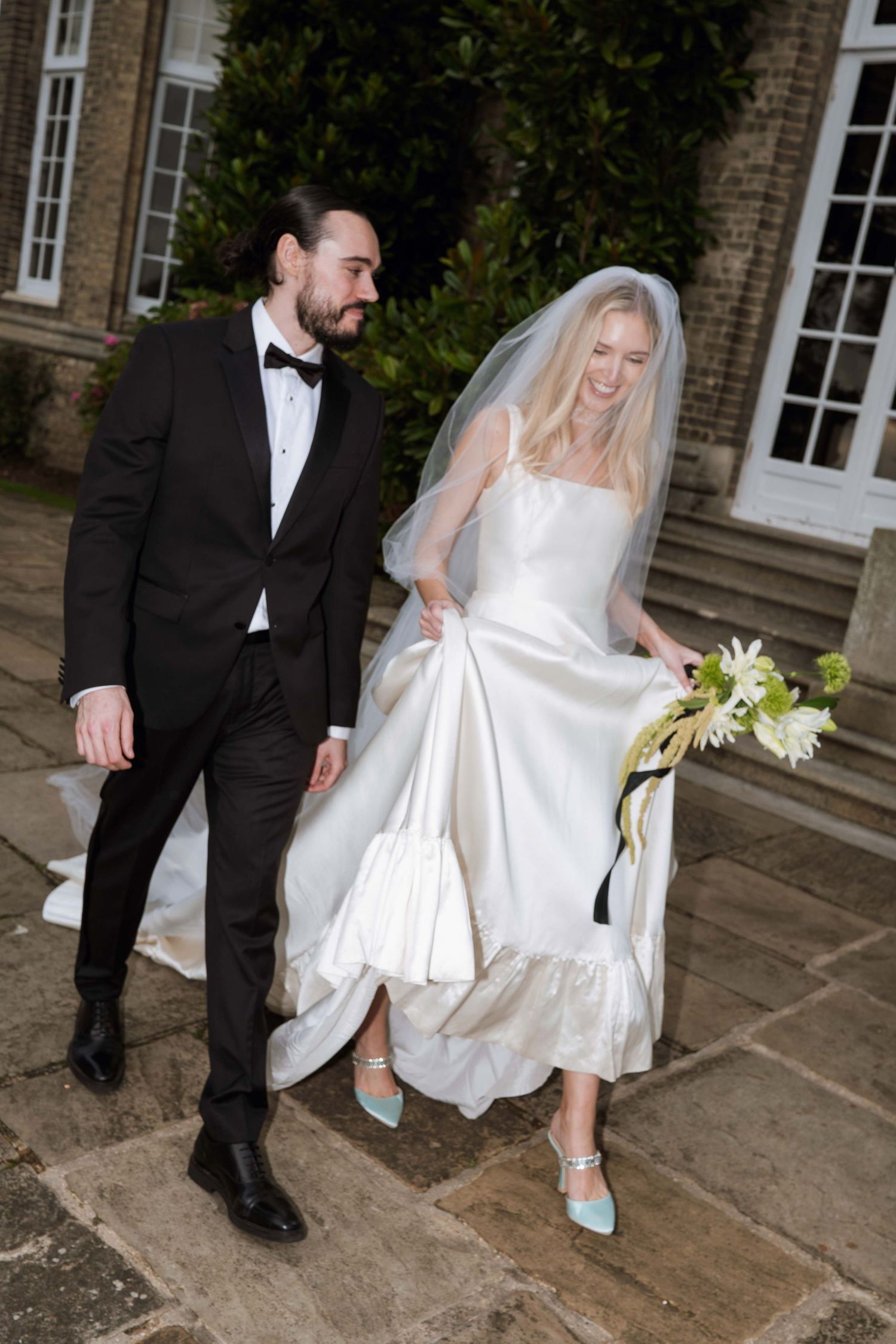 A bride in a white wedding dress and blue shoes walks with a groom in a black tuxedo on stone steps outside a building.