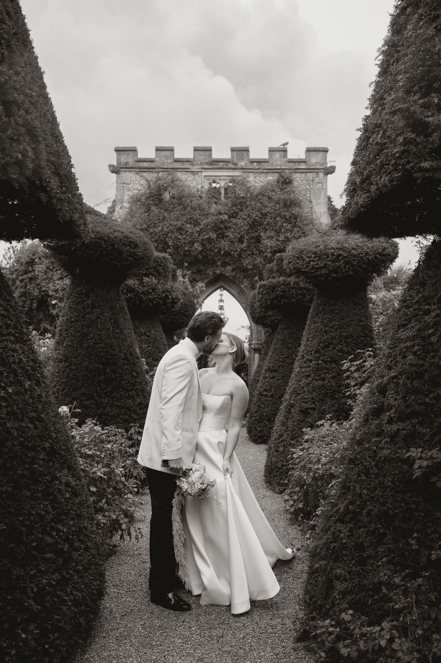 A couple in wedding attire walks through a formal garden path lined with sculpted hedges and trees.