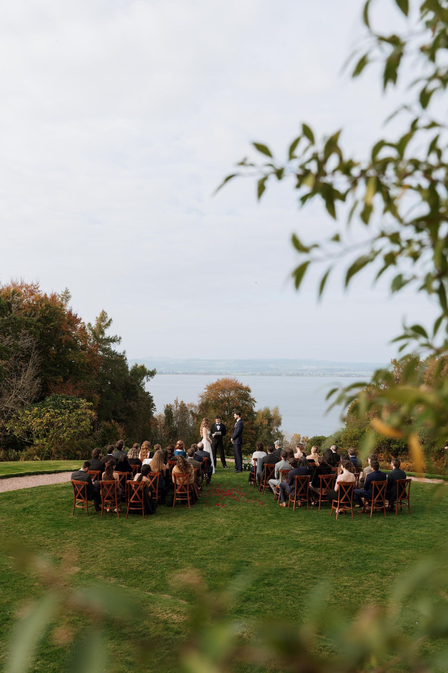 Panoramic view of an outdoor wedding ceremony on a hilltop with guests seated in wooden chairs surrounded by greenery.