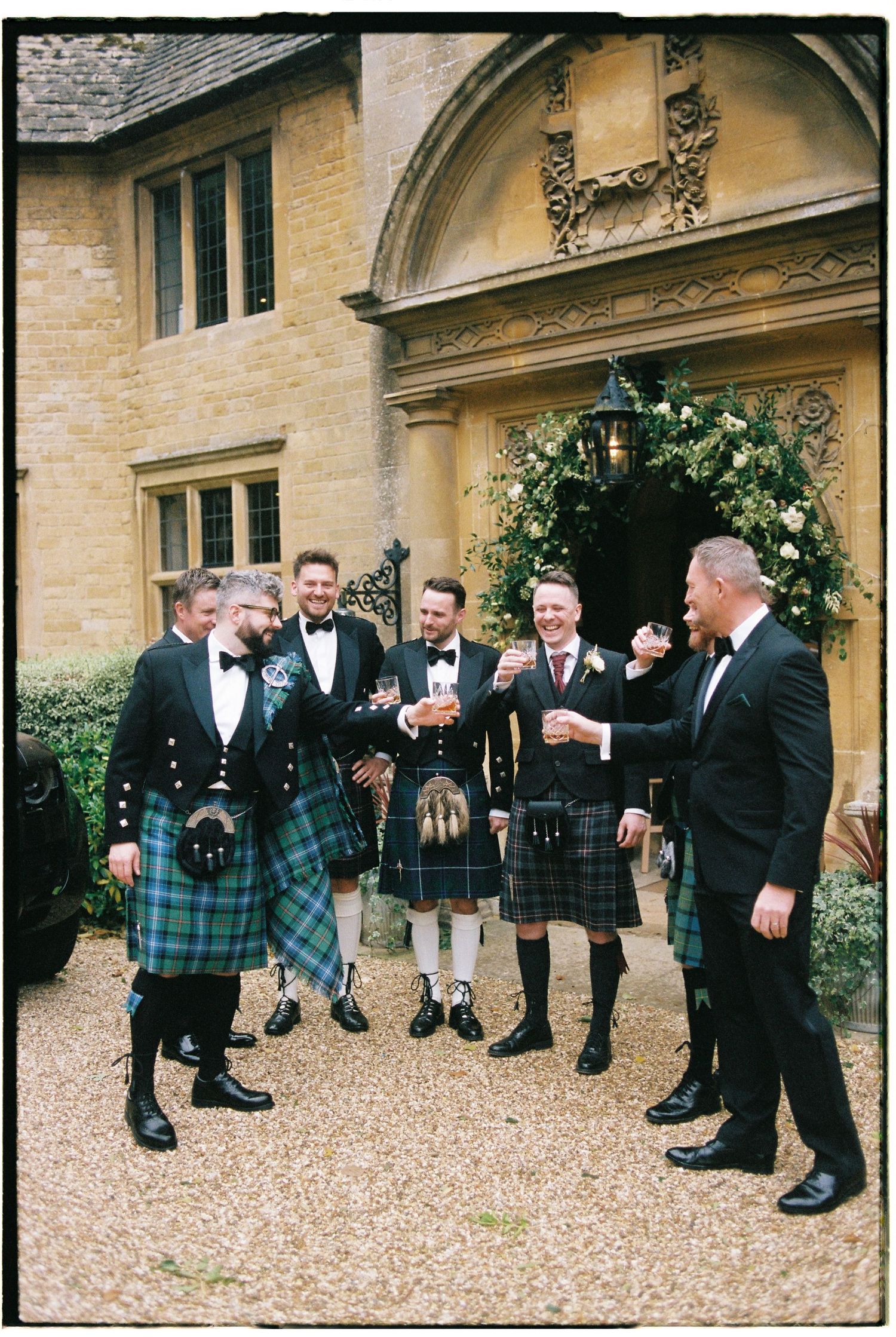 A group of men in formal attire and kilts gather outside a stone building for wedding photos.