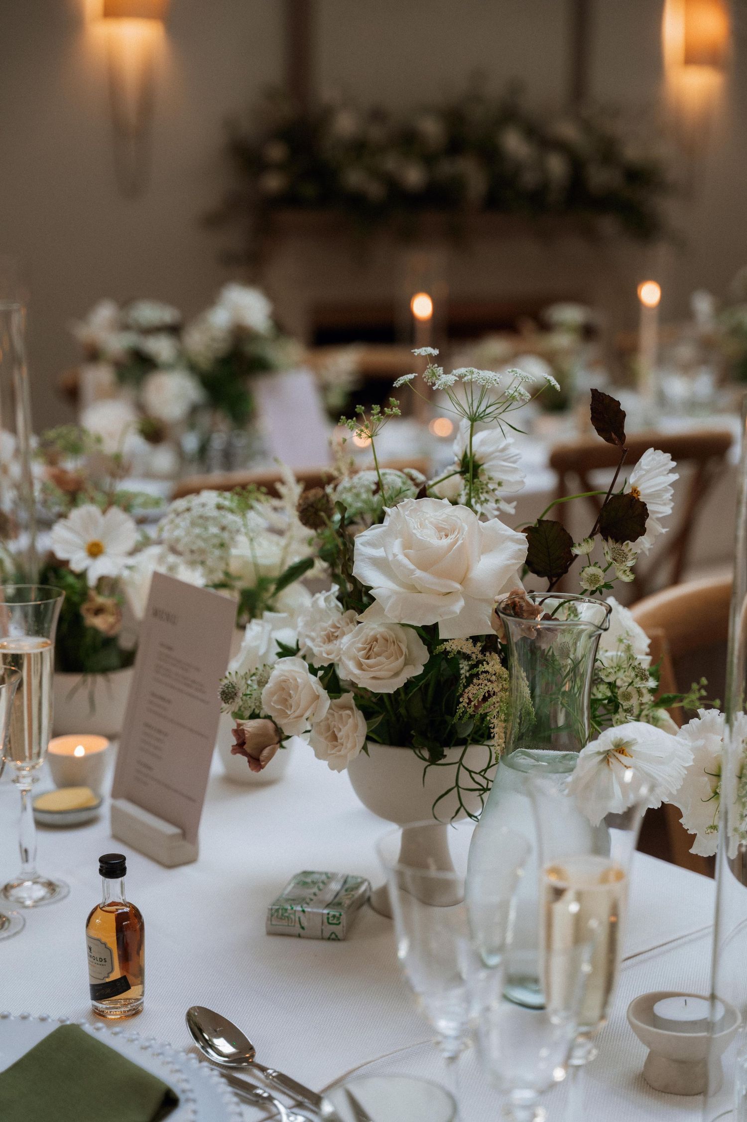 Elegant wedding reception table settings with white roses and candles in glass vases on white tablecloths.
