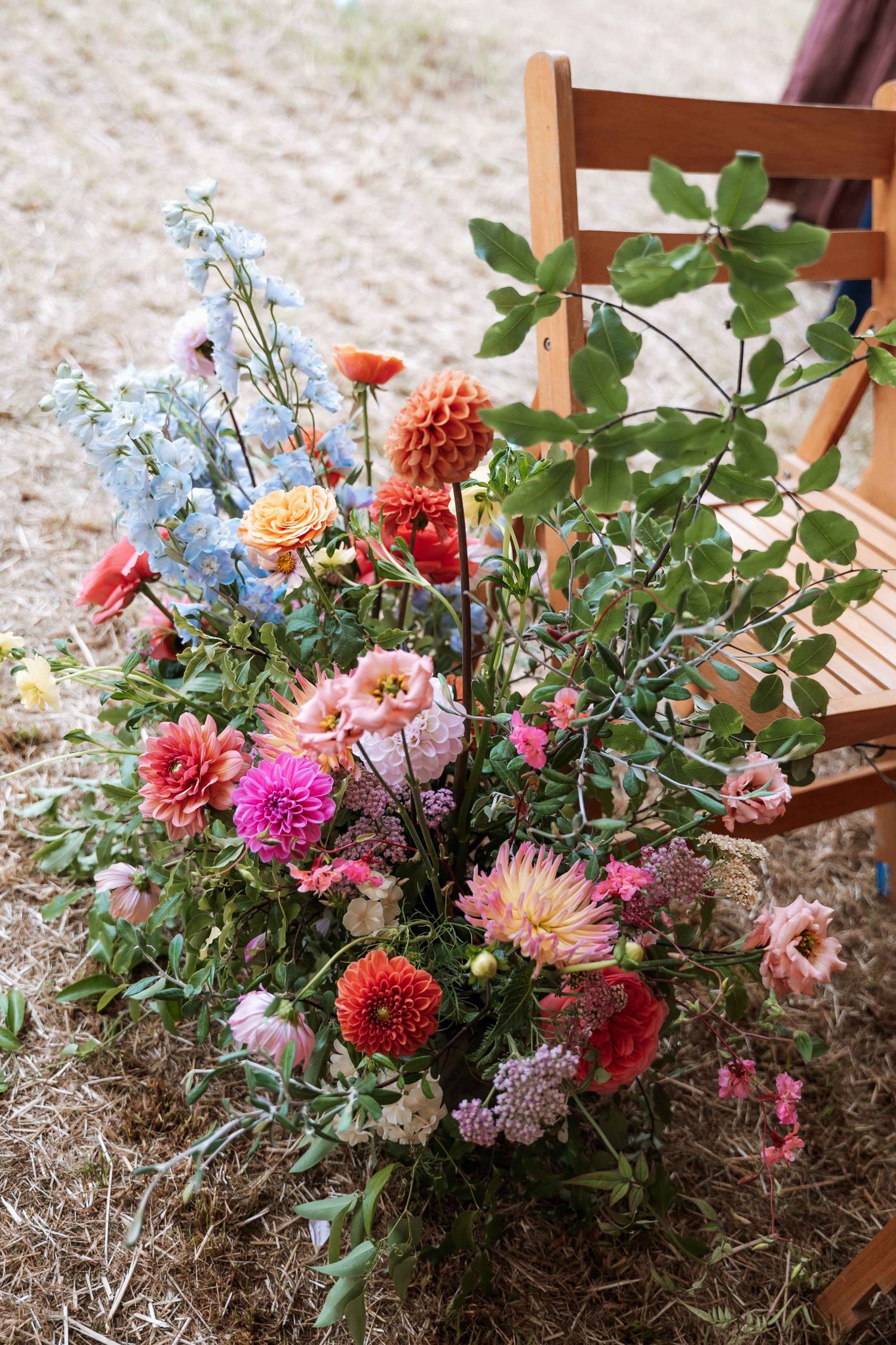 Colorful arrangement of dahlias, delphinium, and garden roses in rustic outdoor floral display on wooden furniture.