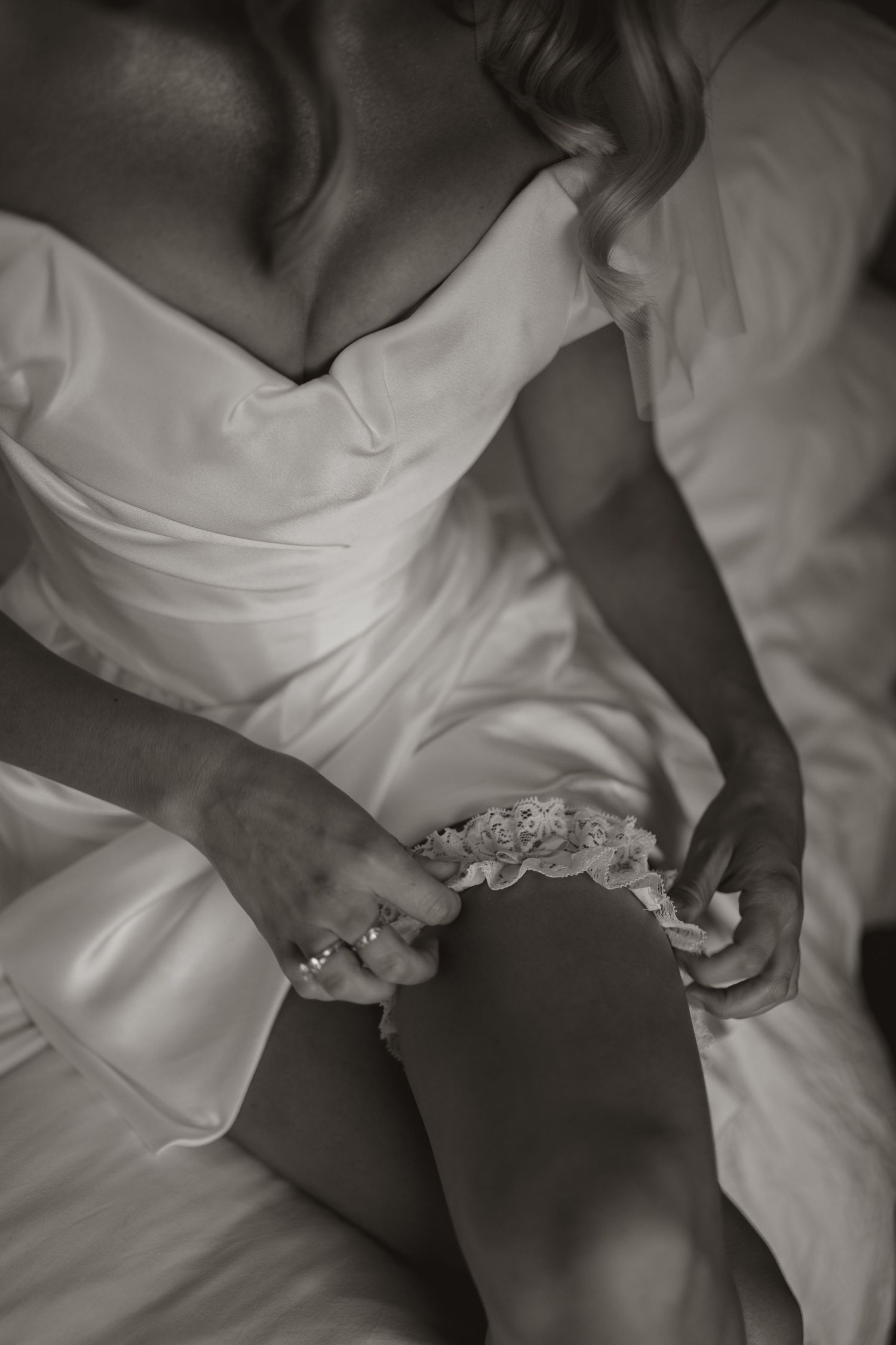 A close-up black and white photograph of a delicate white lace garter on a flowing wedding dress.