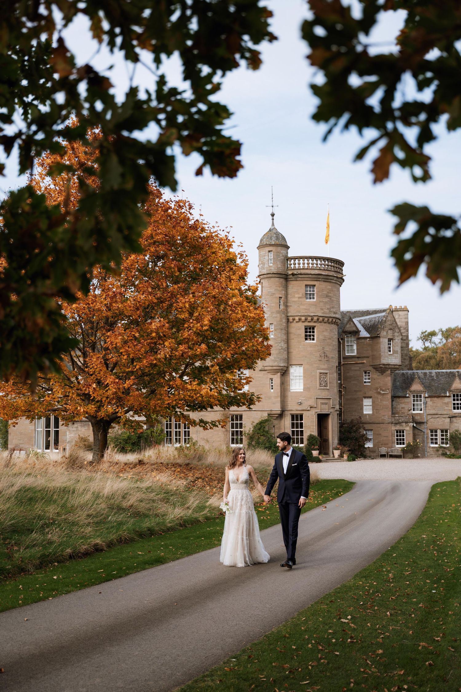 A couple walks along a winding path towards a historic stone castle with turrets on an autumn day in Scotland.
