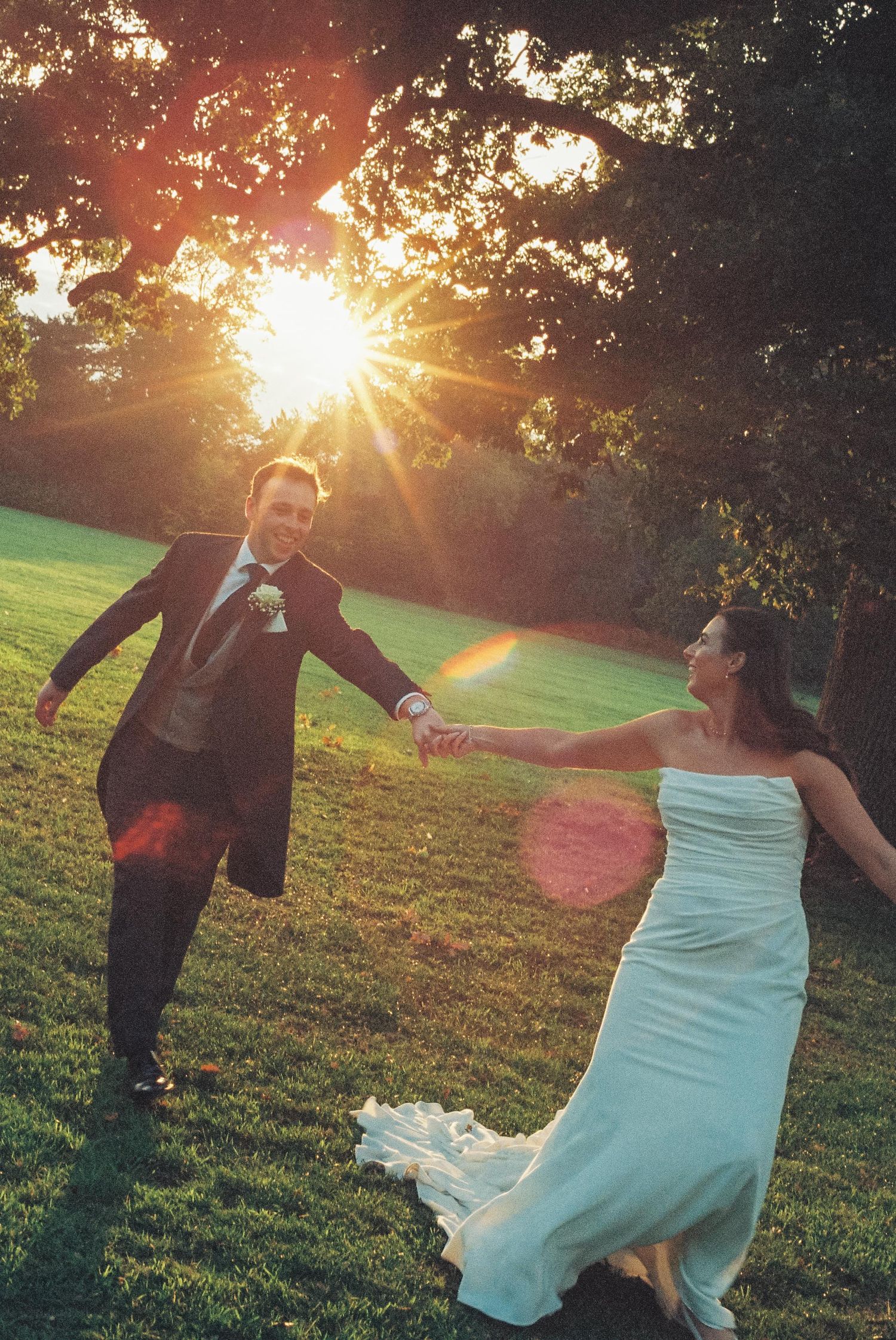 A couple dances together in a grassy field during golden hour sunset with sunbeams streaming through trees.