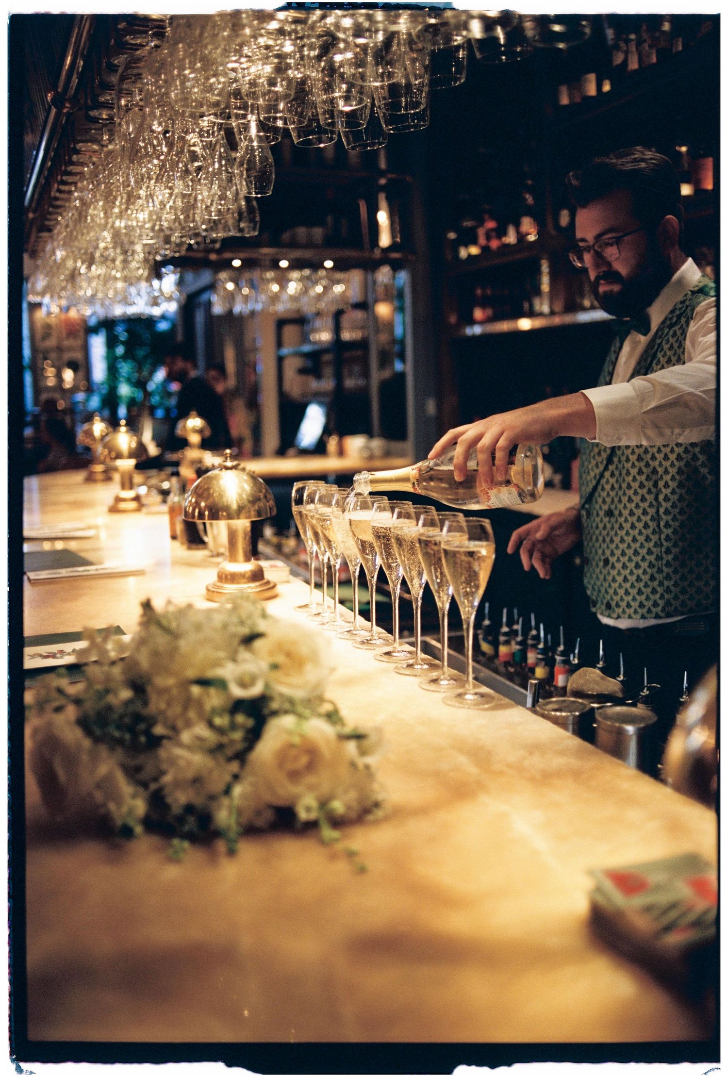 A bartender pours champagne into elegant glasses at an upscale bar with crystal chandeliers and ambient lighting.