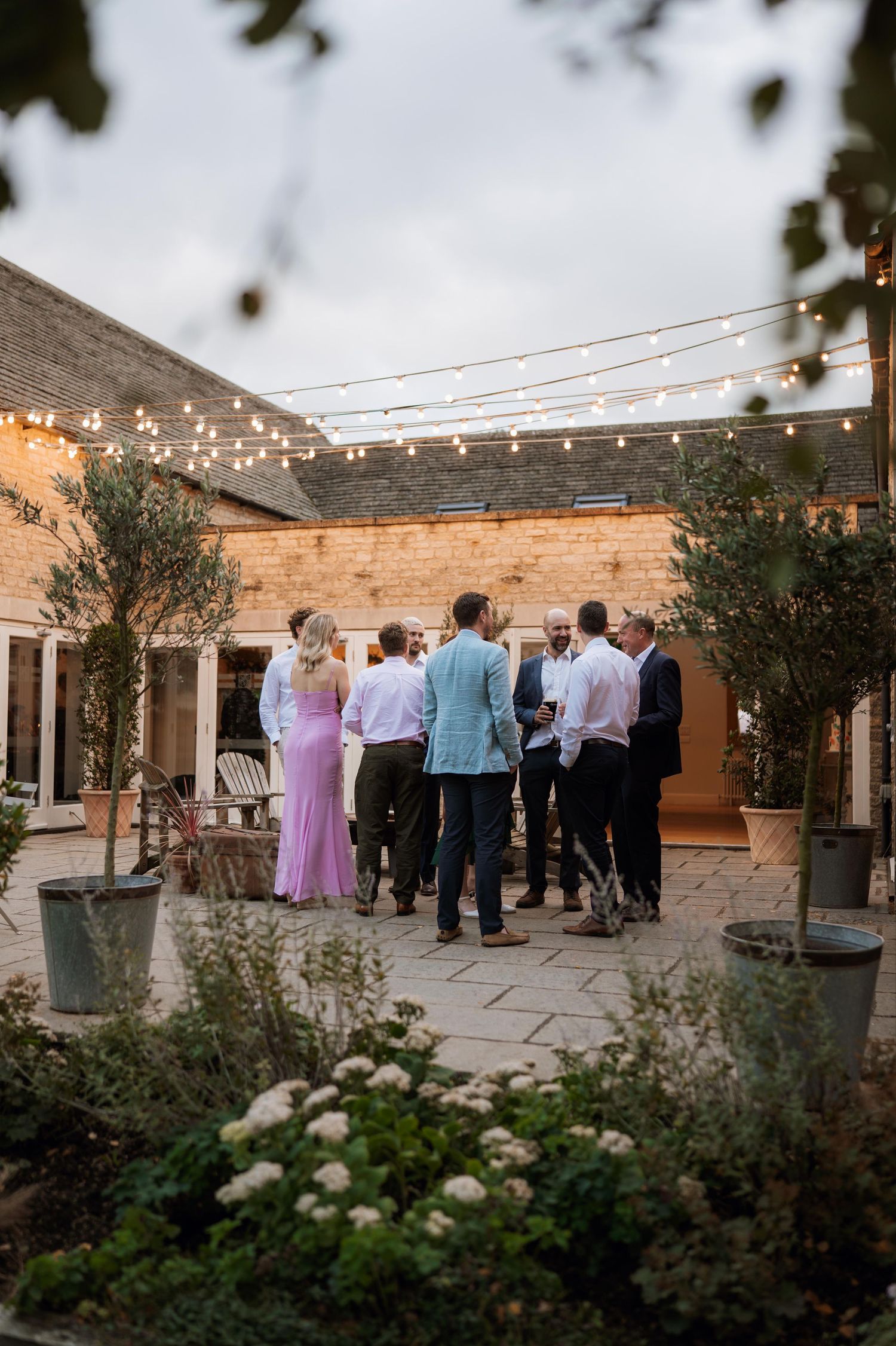 Wedding guests gather on an outdoor patio decorated with string lights and olive trees at dusk.
